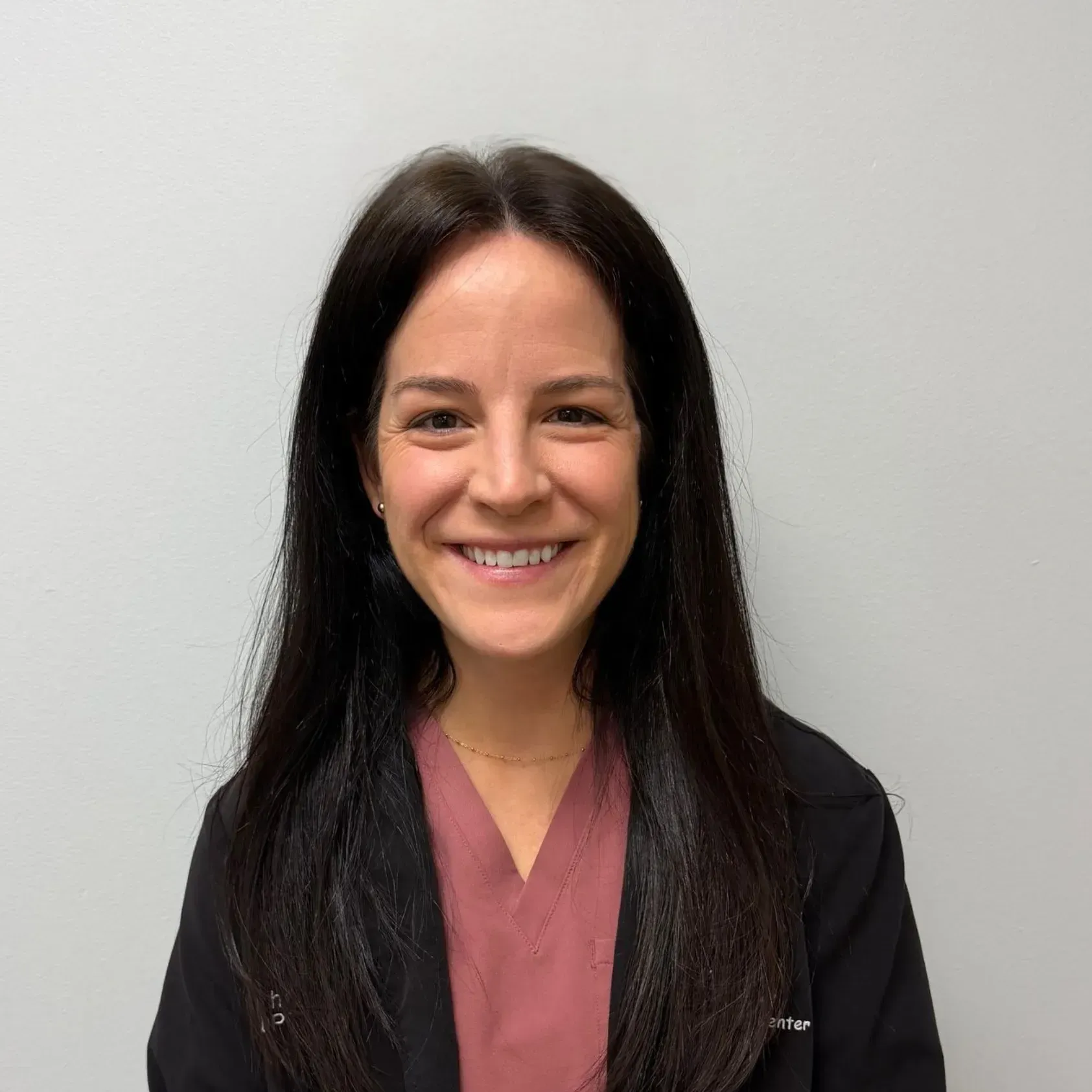 Woman with dark hair smiles, wearing a pink top and black jacket.