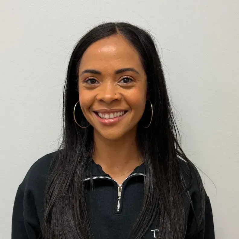 Woman with long dark hair, hoop earrings, and a black sweater smiles at the camera. White background.