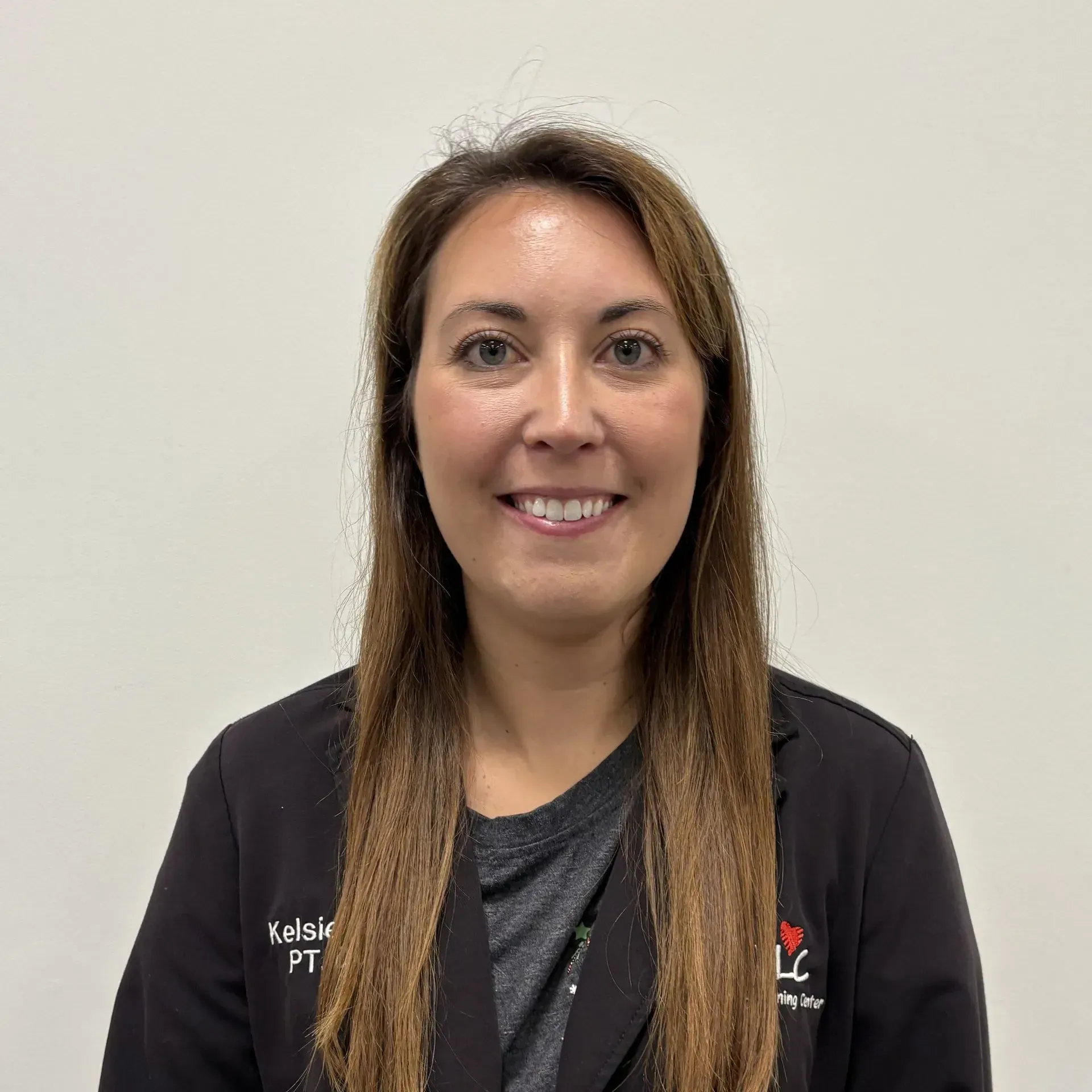 Woman in black jacket smiles, wearing a name tag “Kelsie PT.” White wall backdrop.
