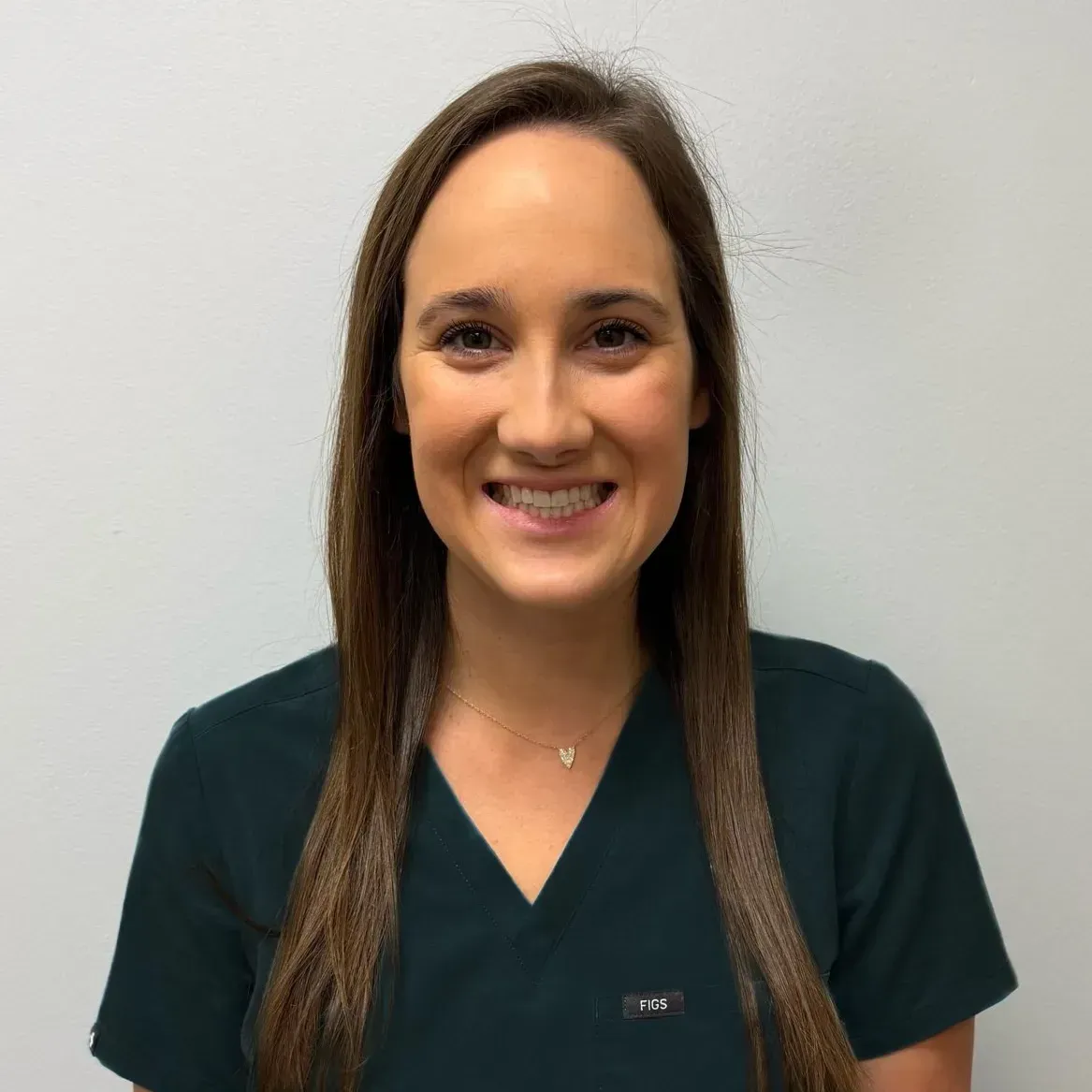 Woman with long brown hair, wearing a dark green scrub top, smiling in front of a white wall.
