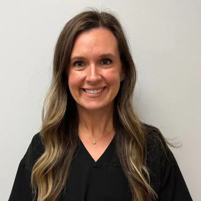 Woman with long brown hair smiling at the camera, wearing black top, gold necklace, white background.