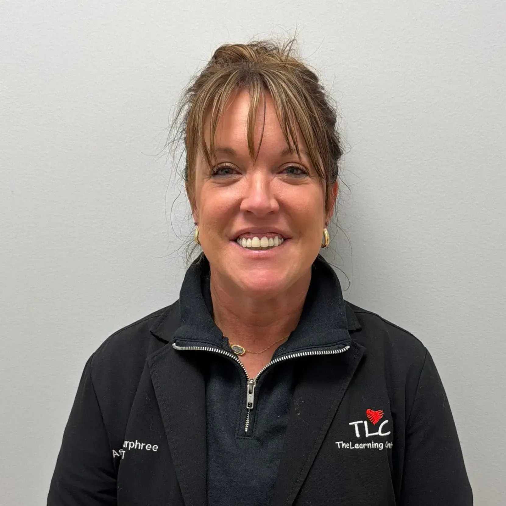 Woman with brown hair smiles, wearing a black jacket with “TLC The Learning Center” logo.
