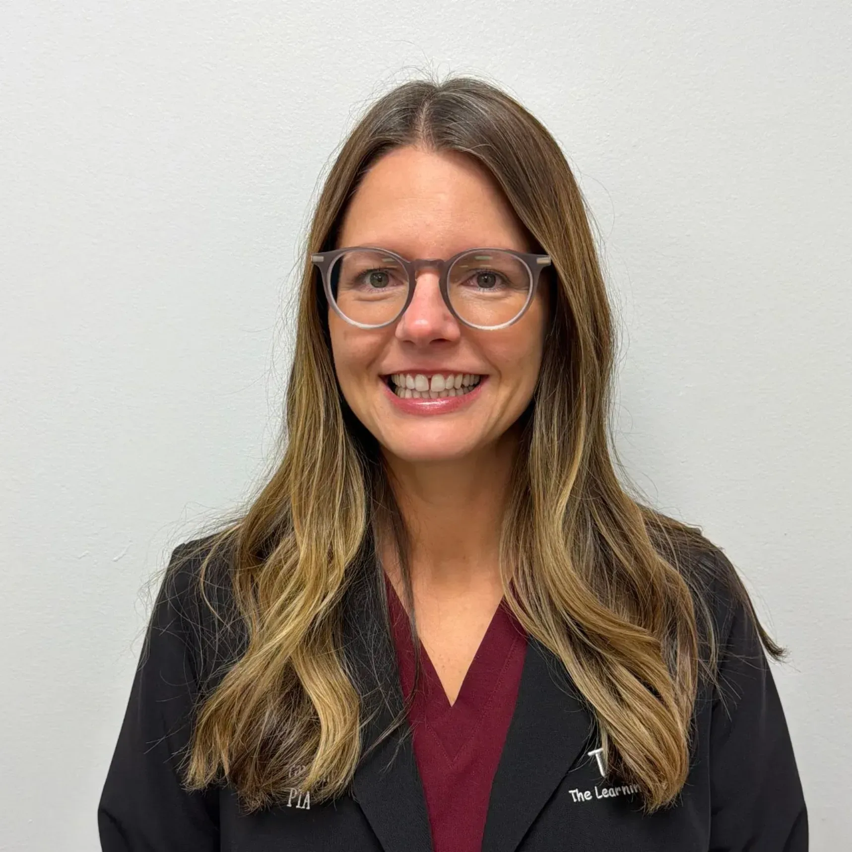 Woman wearing glasses, a burgundy top, and a black coat smiles in front of a white background.