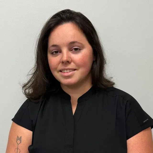 Woman with dark hair smiles, wearing a black shirt, and stands in front of a white wall.