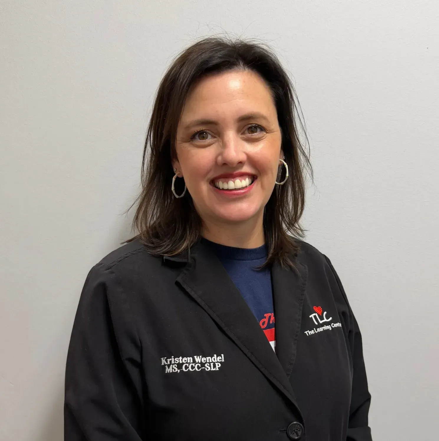Woman with brown hair, smiling, wearing black jacket, silver hoop earrings, and name tag.
