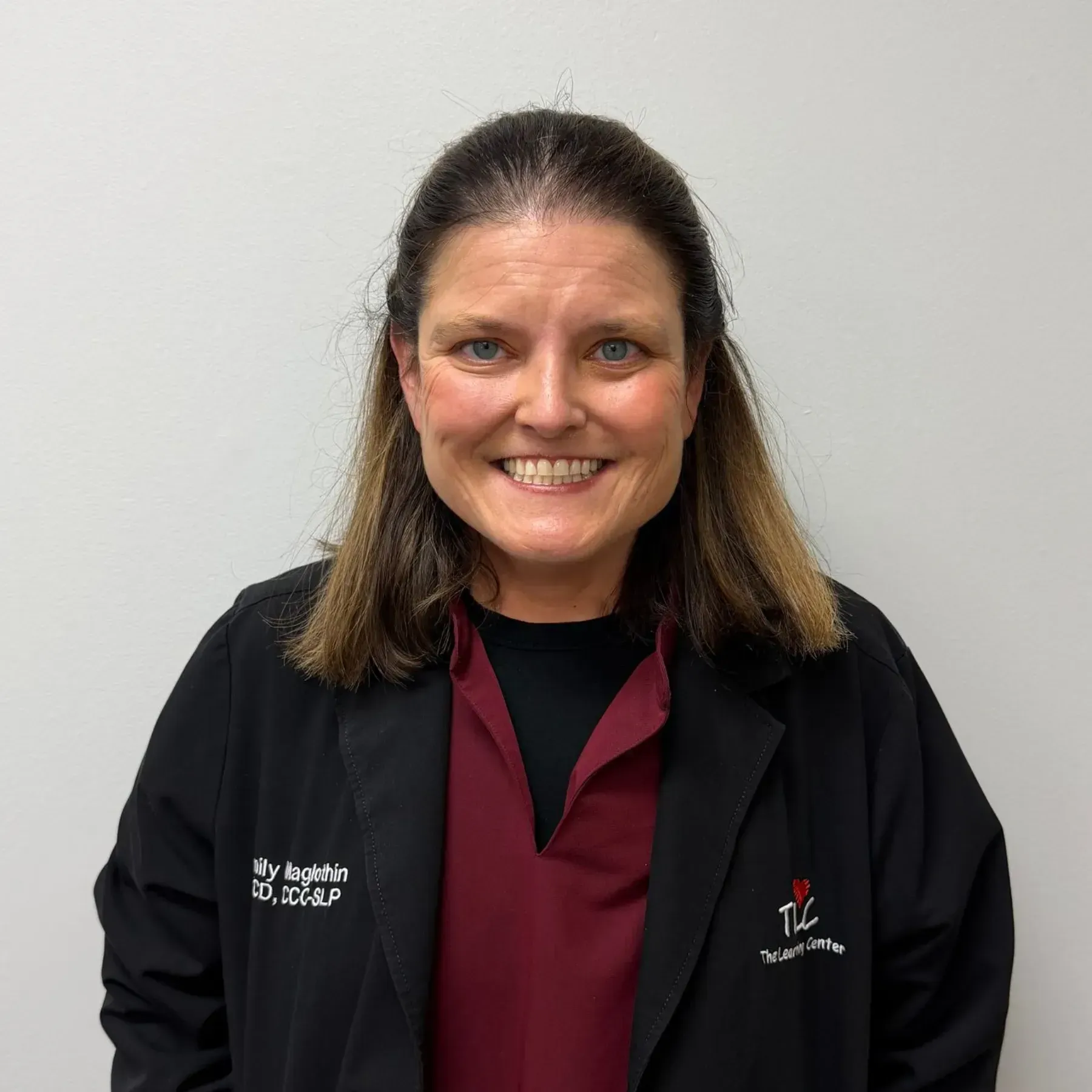 Smiling woman in black lab coat, maroon shirt, TLC logo, against a white wall.