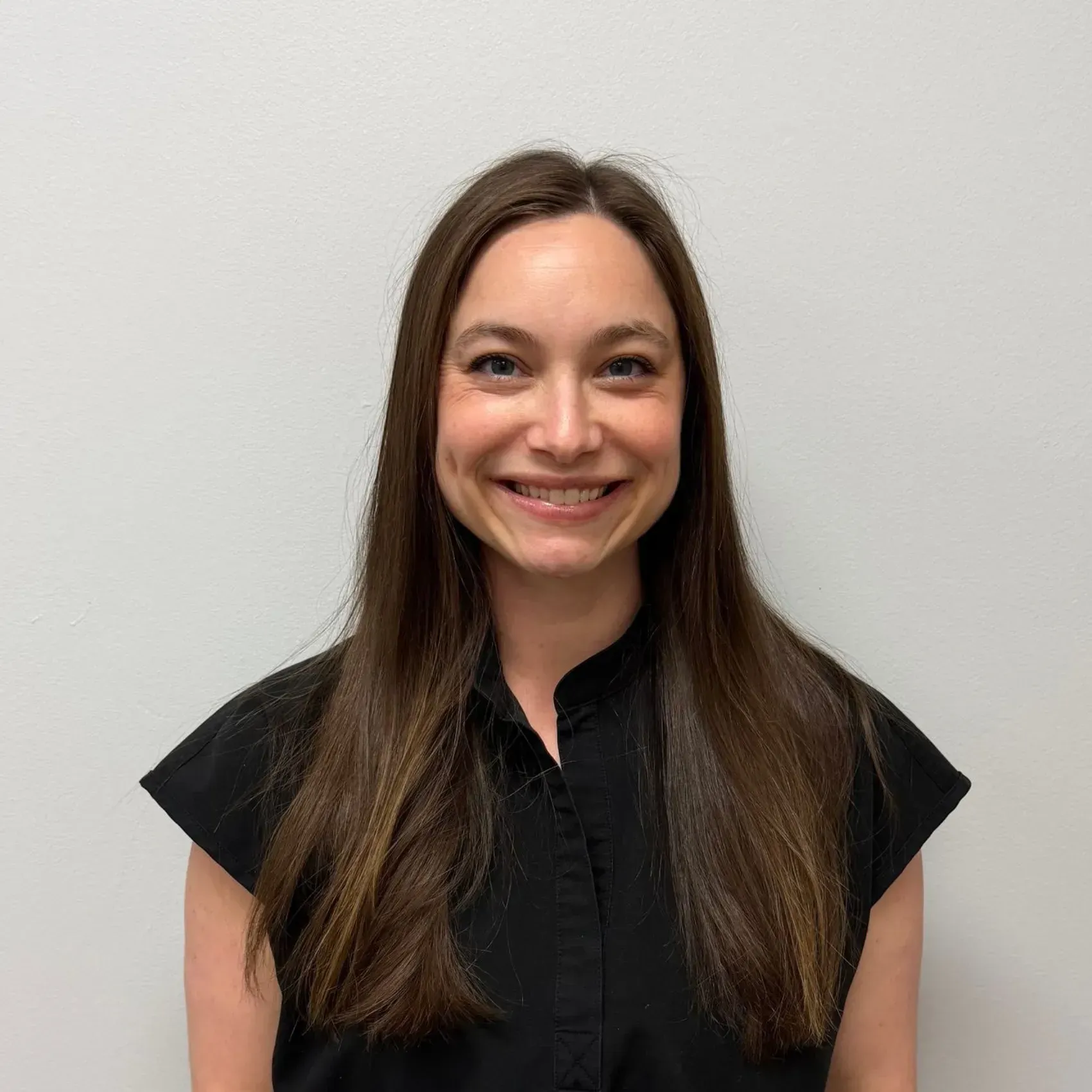 Woman with long brown hair smiles, wearing a black top against a white wall.