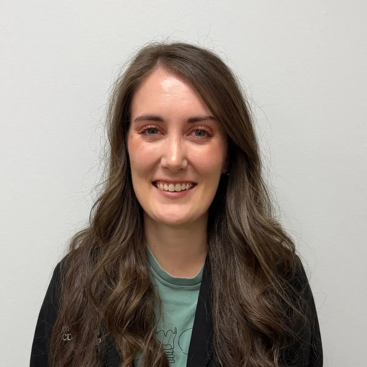 Woman with long brown hair smiles, wearing a green shirt and black jacket.