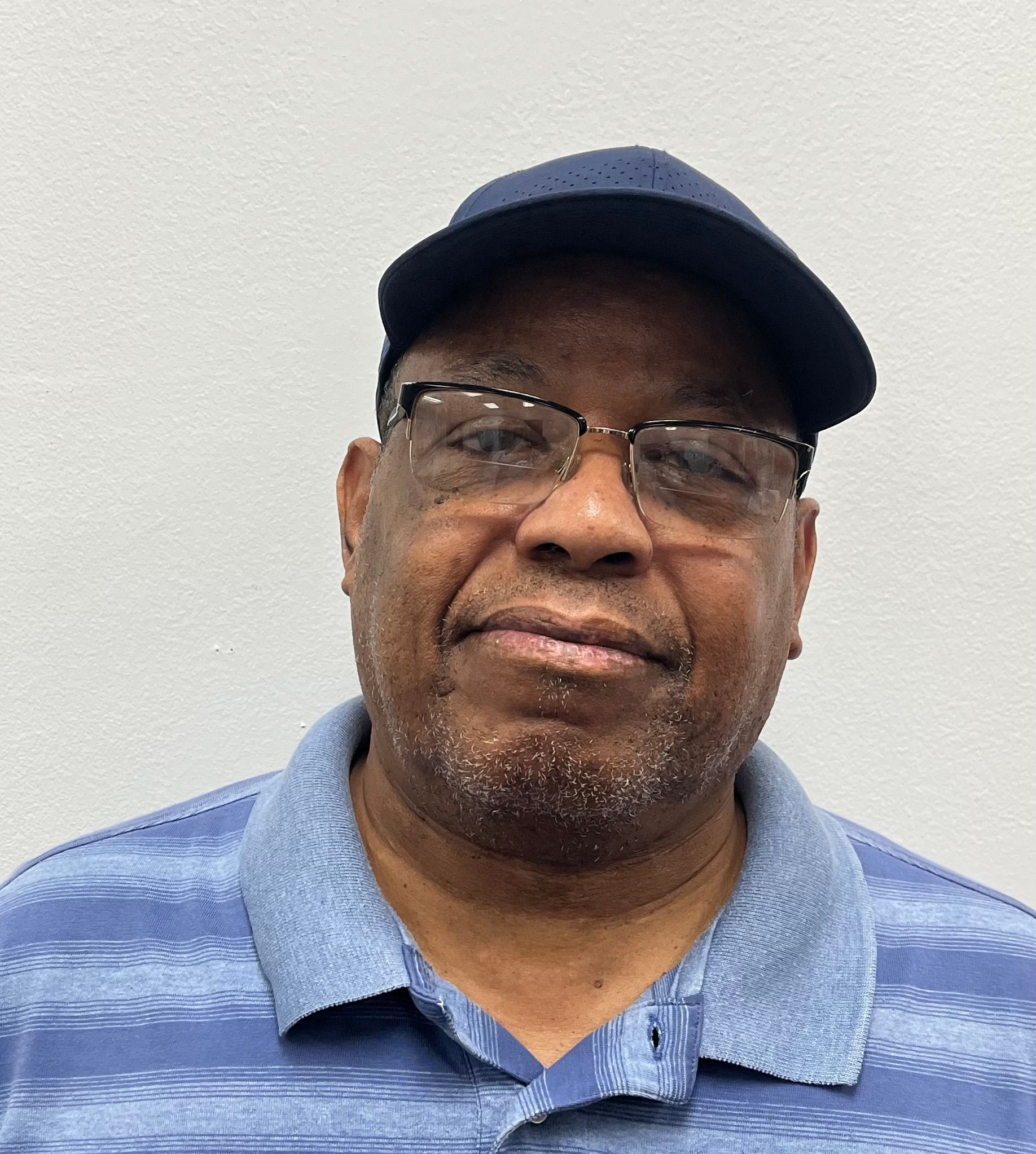 Man wearing glasses and a navy cap, smiling slightly against a plain light wall.