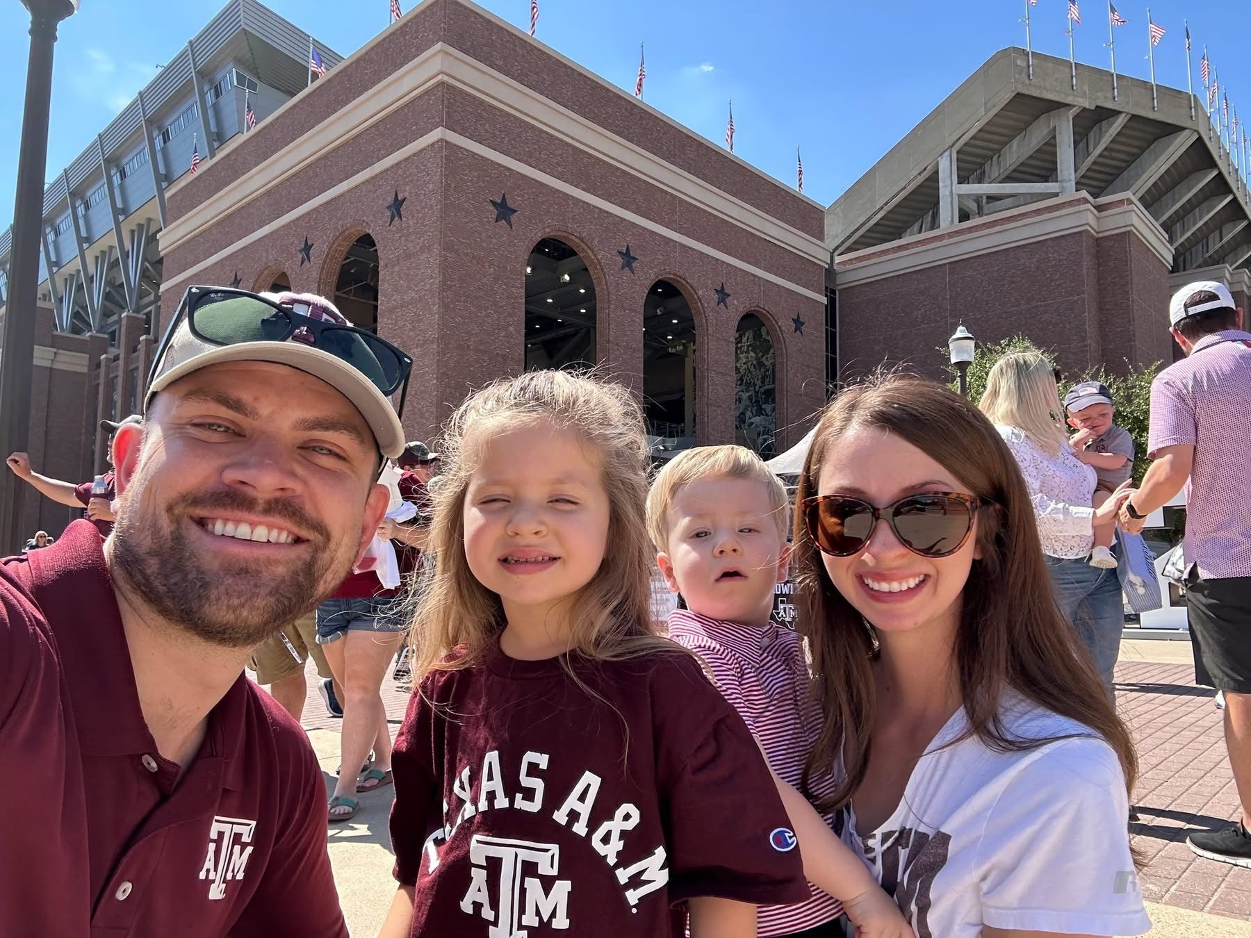 Family selfie in front of a brick stadium. People wearing maroon and white, smiling.