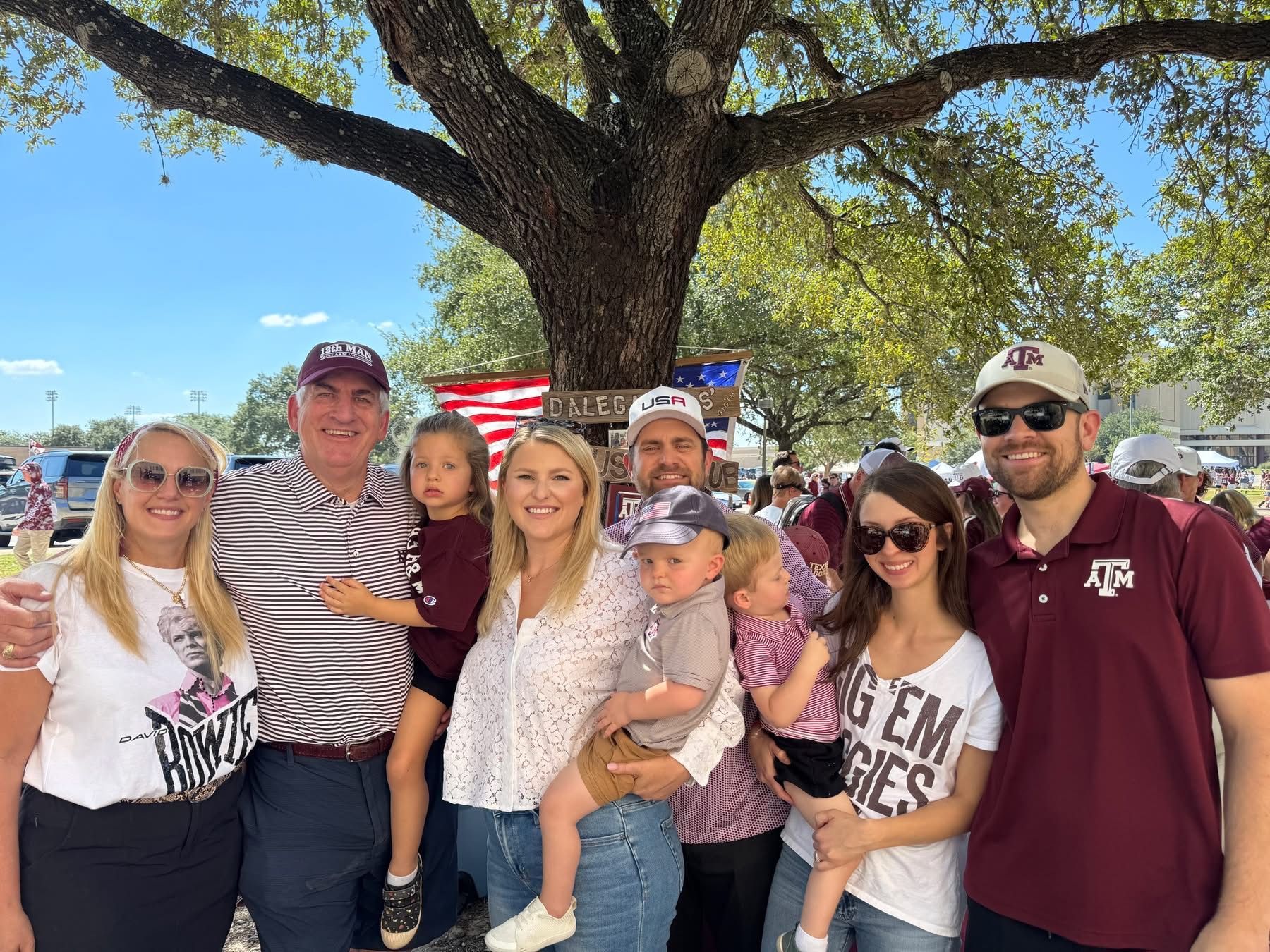Group of people at an outdoor gathering, wearing maroon and white; trees and an American flag in the background.