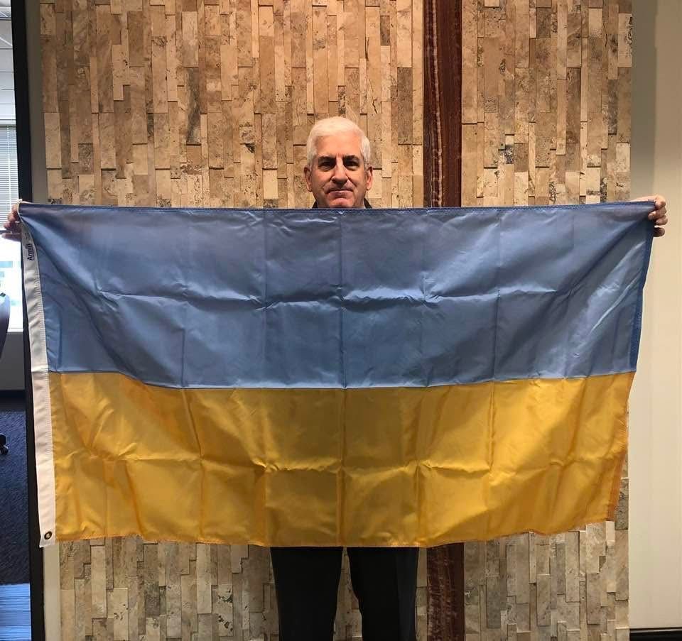 Man holding a Ukrainian flag against a textured wall; flag is blue and yellow.