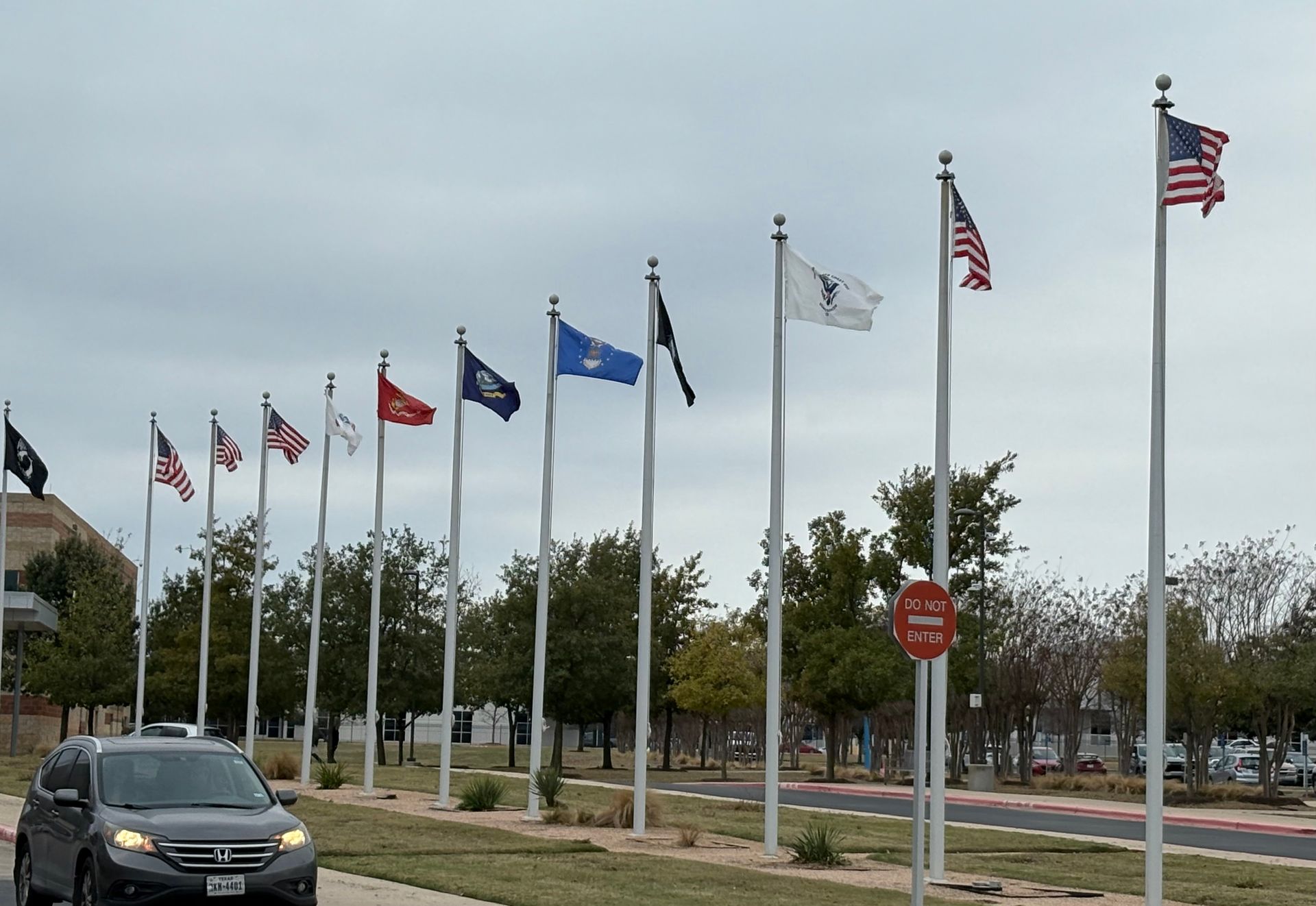 Flags on tall poles, including American flag, line a street; a vehicle drives past.