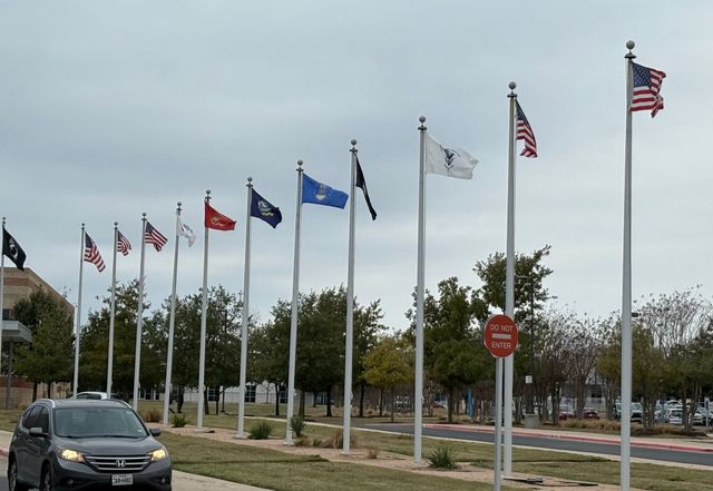Row of flags on poles, including US flags, with a vehicle driving past.