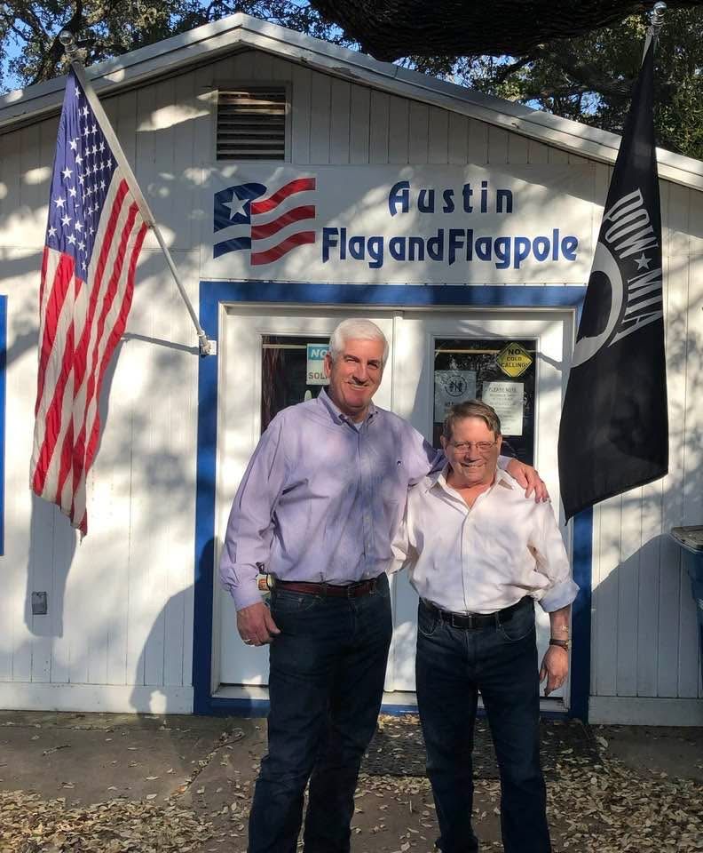 Two men standing in front of Austin Flag and Flagpole store with US and POW flags.