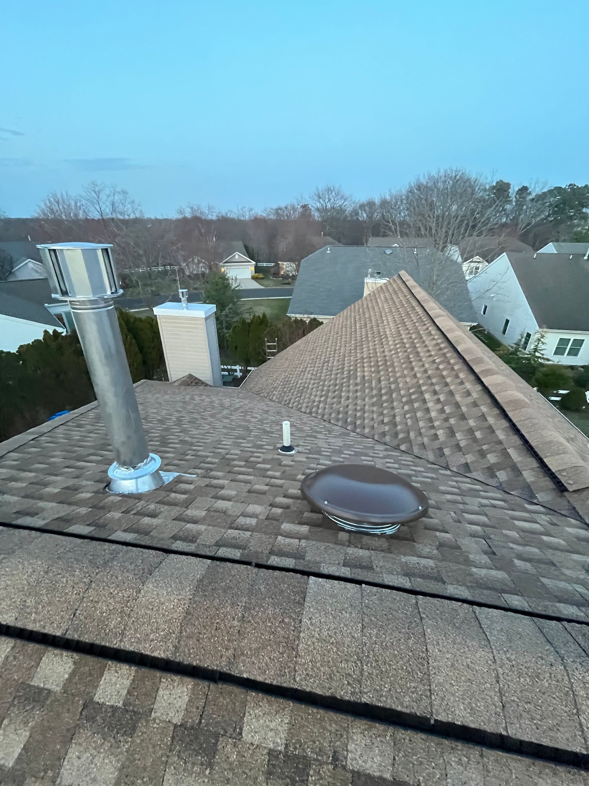A view from a roof showing a metal chimney pipe, a small white vent pipe, and a dome-shaped skylight against a blue sky.