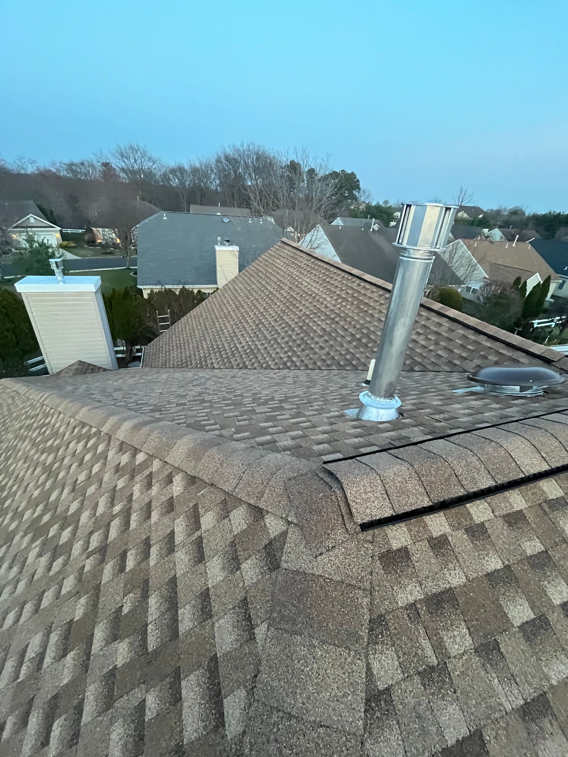 A high-angle view of a brown shingled roof with a metal ventilation pipe and a small chimney against a blue sky.