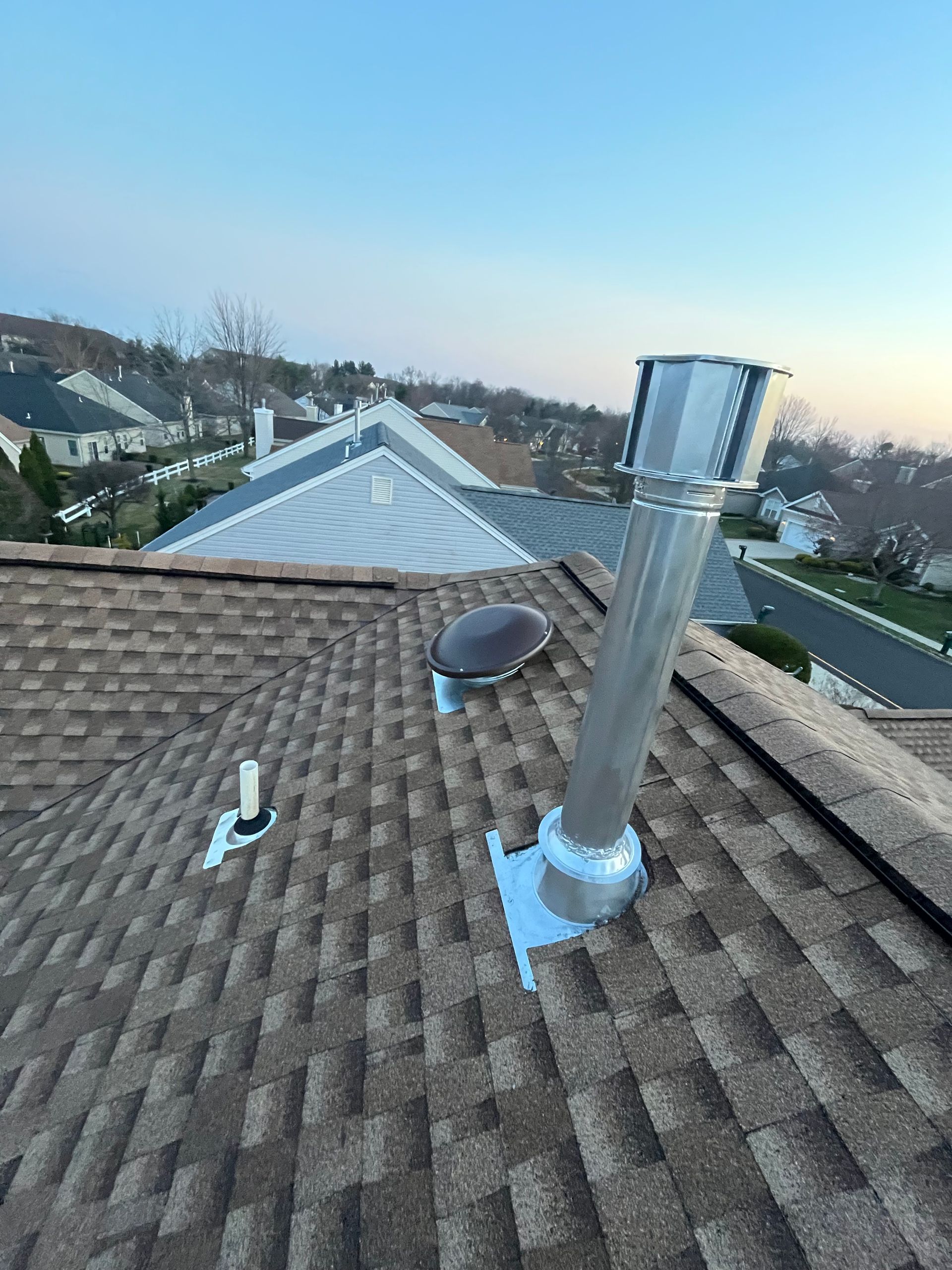 A metal chimney pipe, a circular roof vent, and a small white plumbing vent on a shingled roof under a clear blue sky.