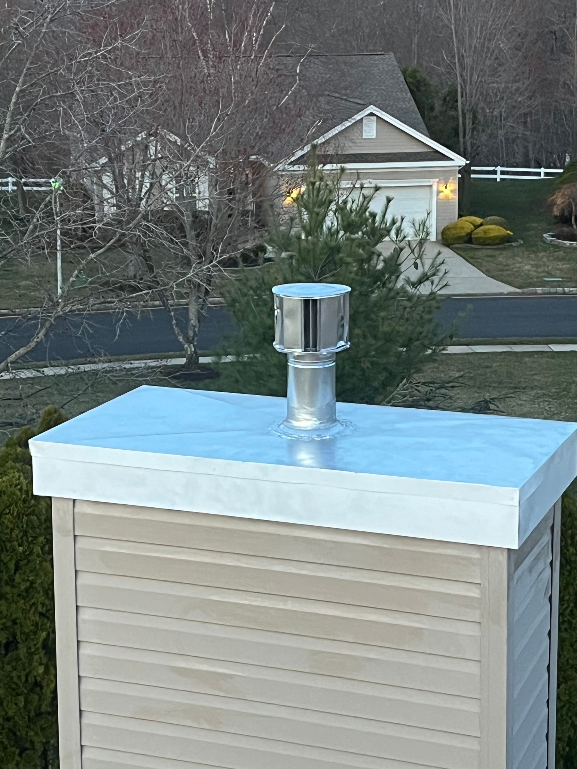 A metal chimney cap sits on a beige, vinyl-sided chimney chase, with a suburban house and trees in the background.