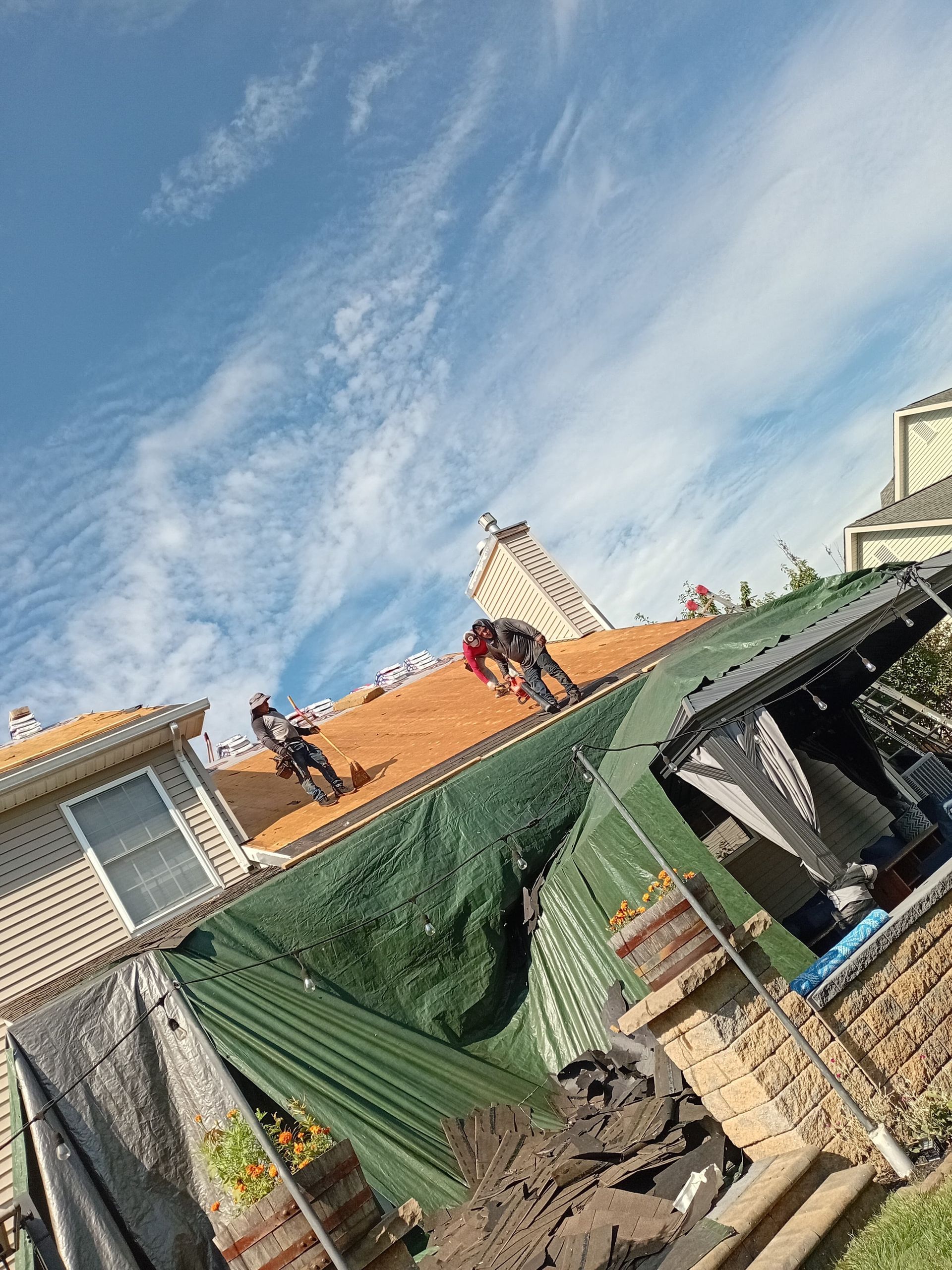 Tilted view of rooftops under a blue sky, with workers on a house roof and a green tarp.