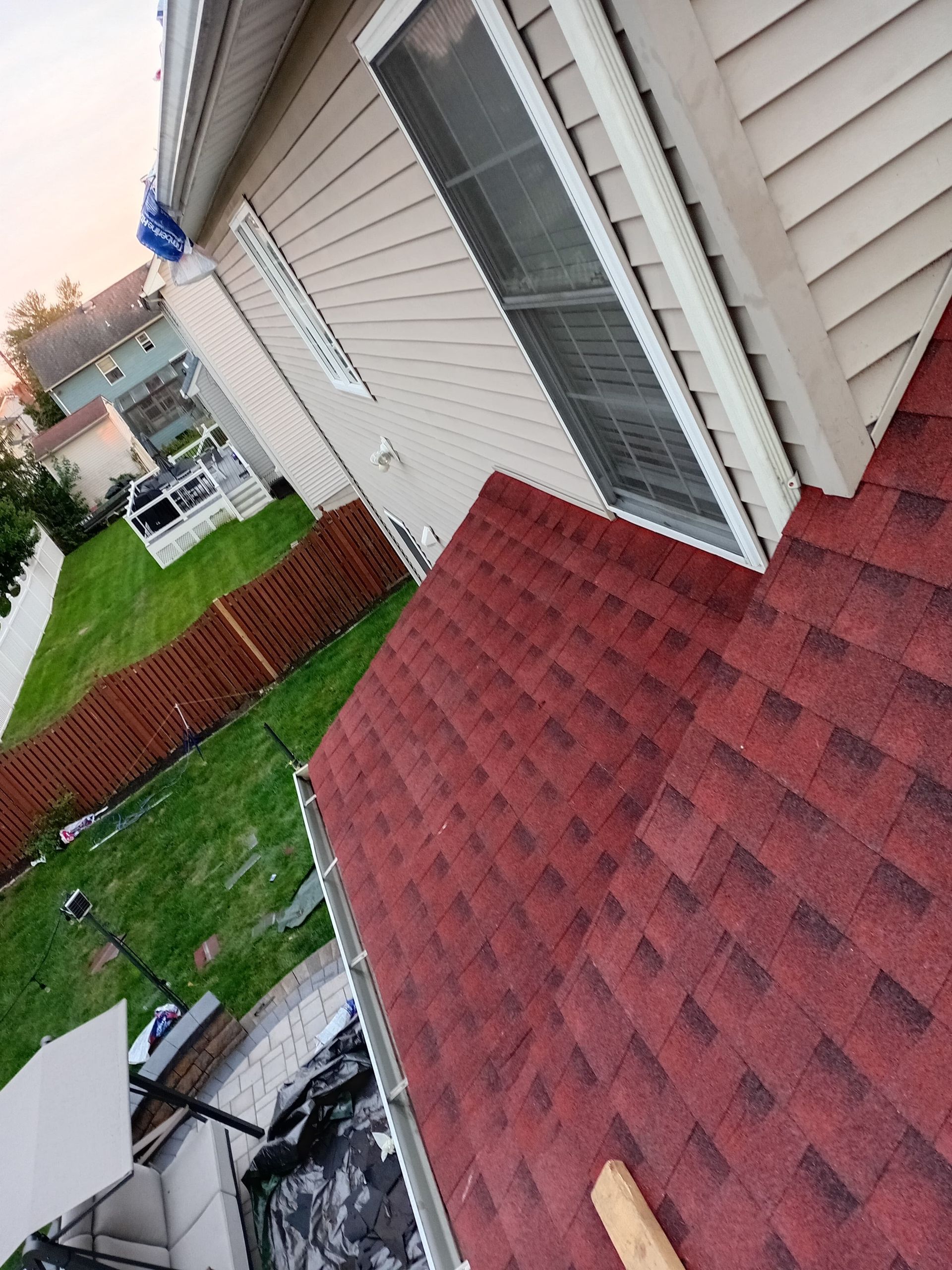 Tilted view of a house roof with red shingles beside beige siding, a window, and a fenced backyard lawn.