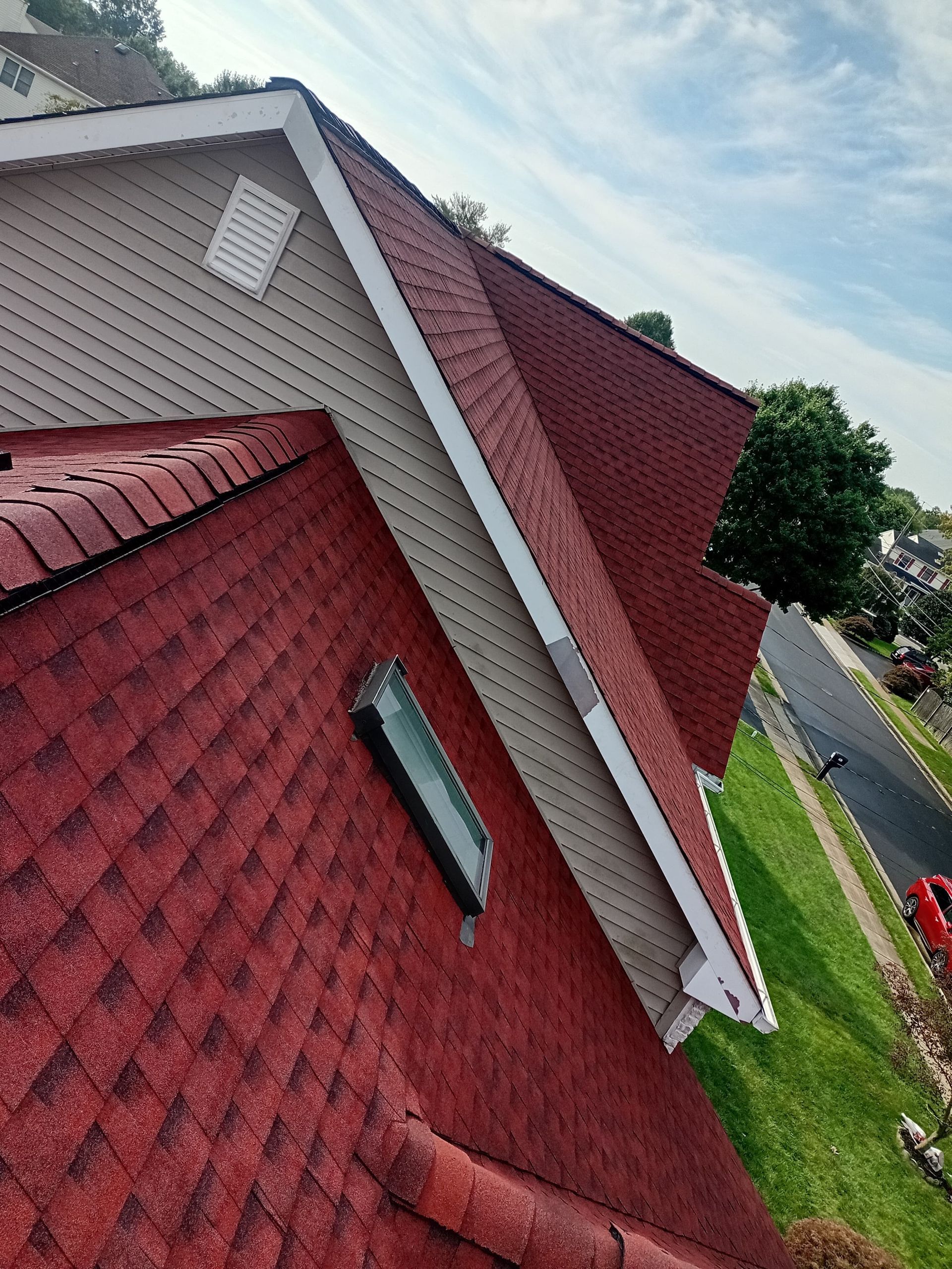Tilted view of red shingle roof and white siding house beside a grassy street under cloudy sky