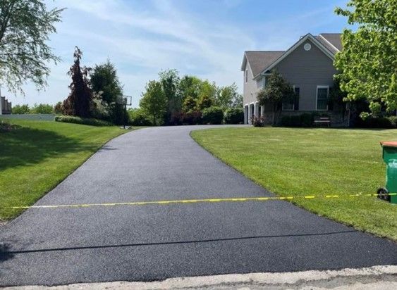 Freshly paved black asphalt driveway leading up to a two-story house with a green lawn under a blue sky.