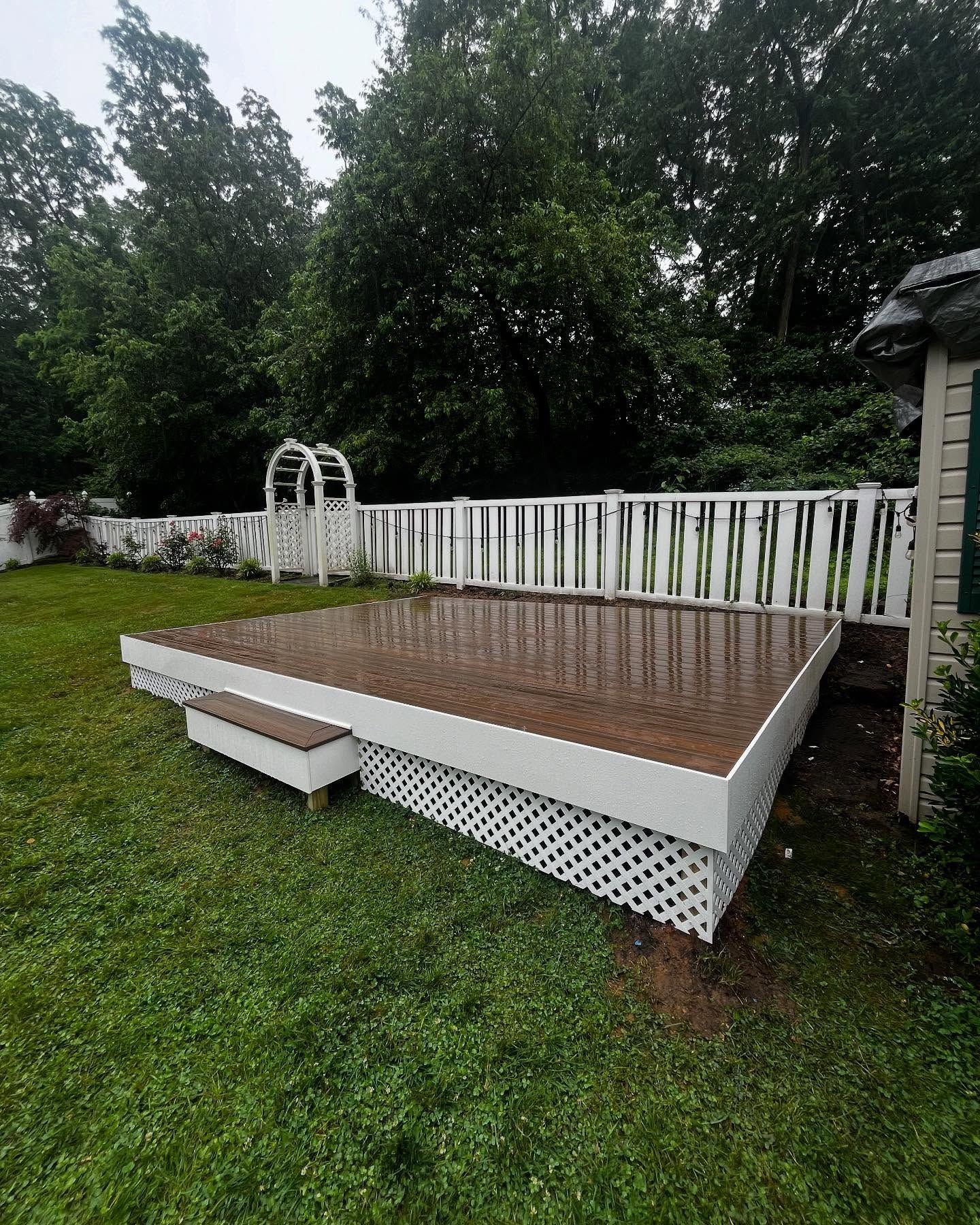 White deck with lattice skirting in a green yard, near a picket fence and trees.