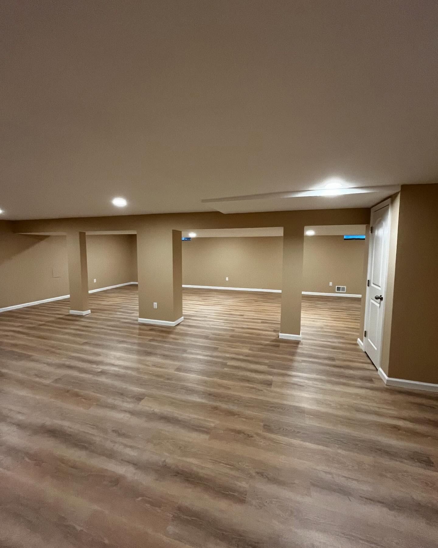 Finished basement with wood-look flooring, beige walls and support beams, and a white door.