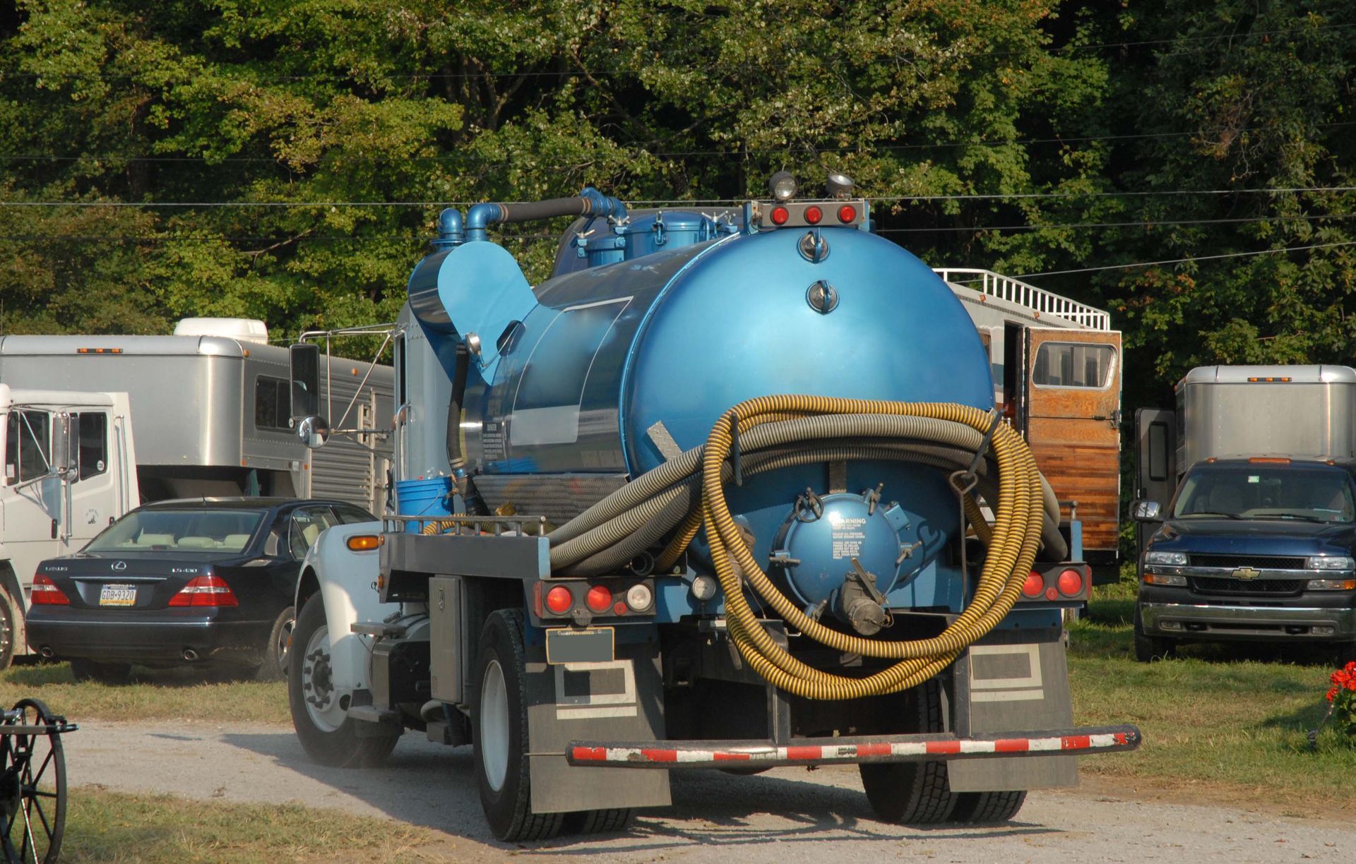 Blue utility truck with a large cylindrical tank and coiled hose parked outdoors among trailers and cars