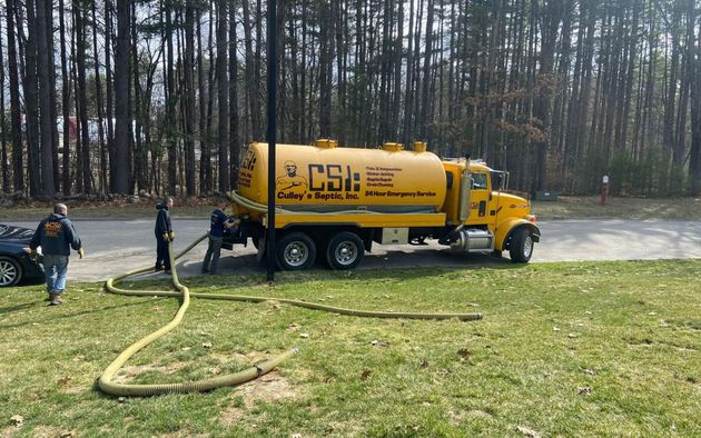 Yellow tanker truck on grass beside road, with workers handling hoses near a wooded area.