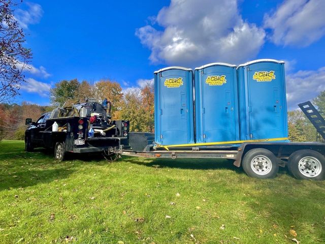 Black truck towing three blue portable toilets on a trailer in a grassy field under a bright blue sky