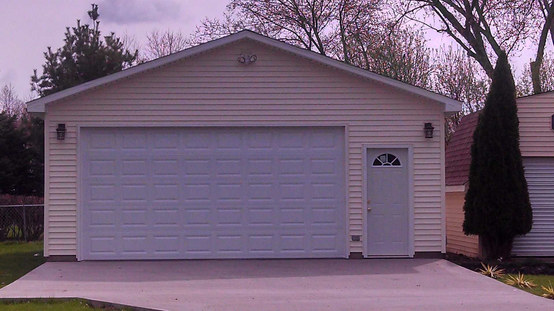 Single-story beige garage with a white door and side entrance, set beside a driveway and trees.