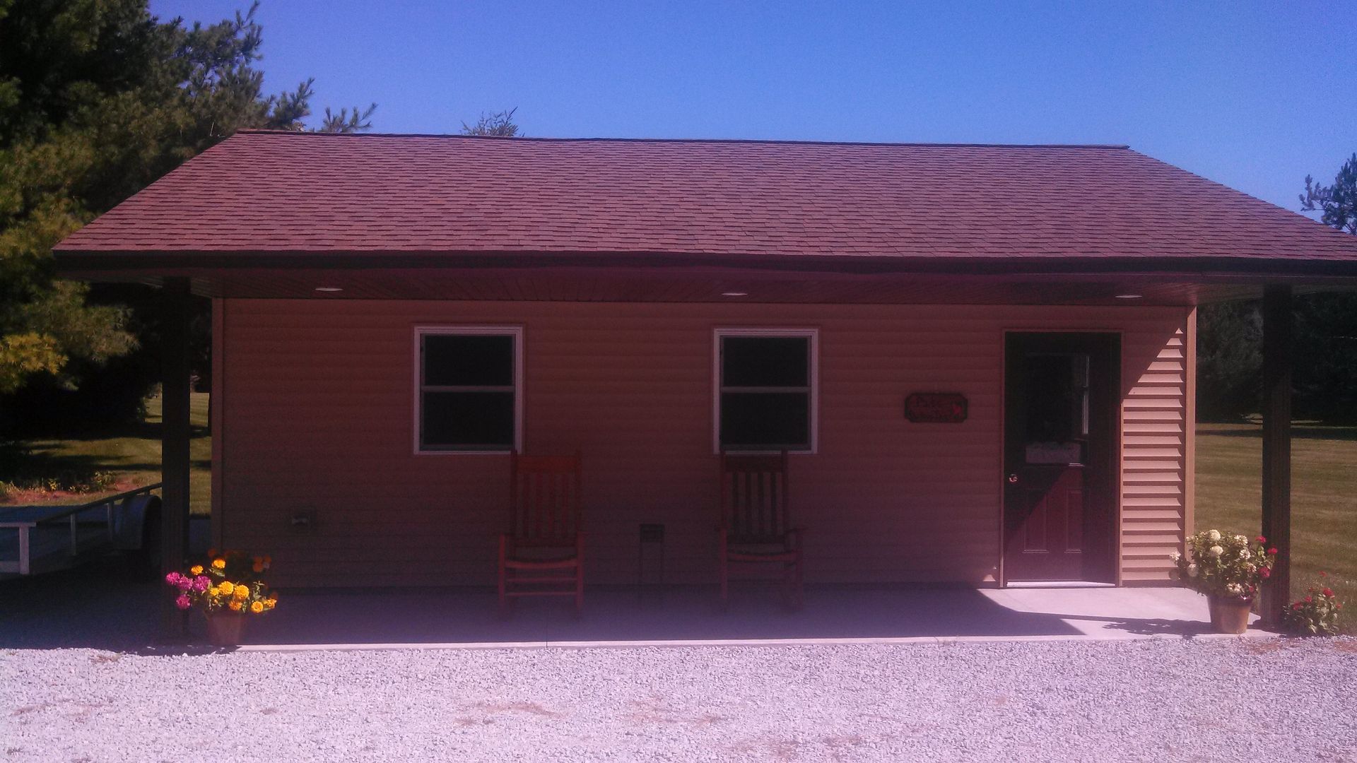 Single-story red house with a front porch, gravel yard, and flower pots in sunlight