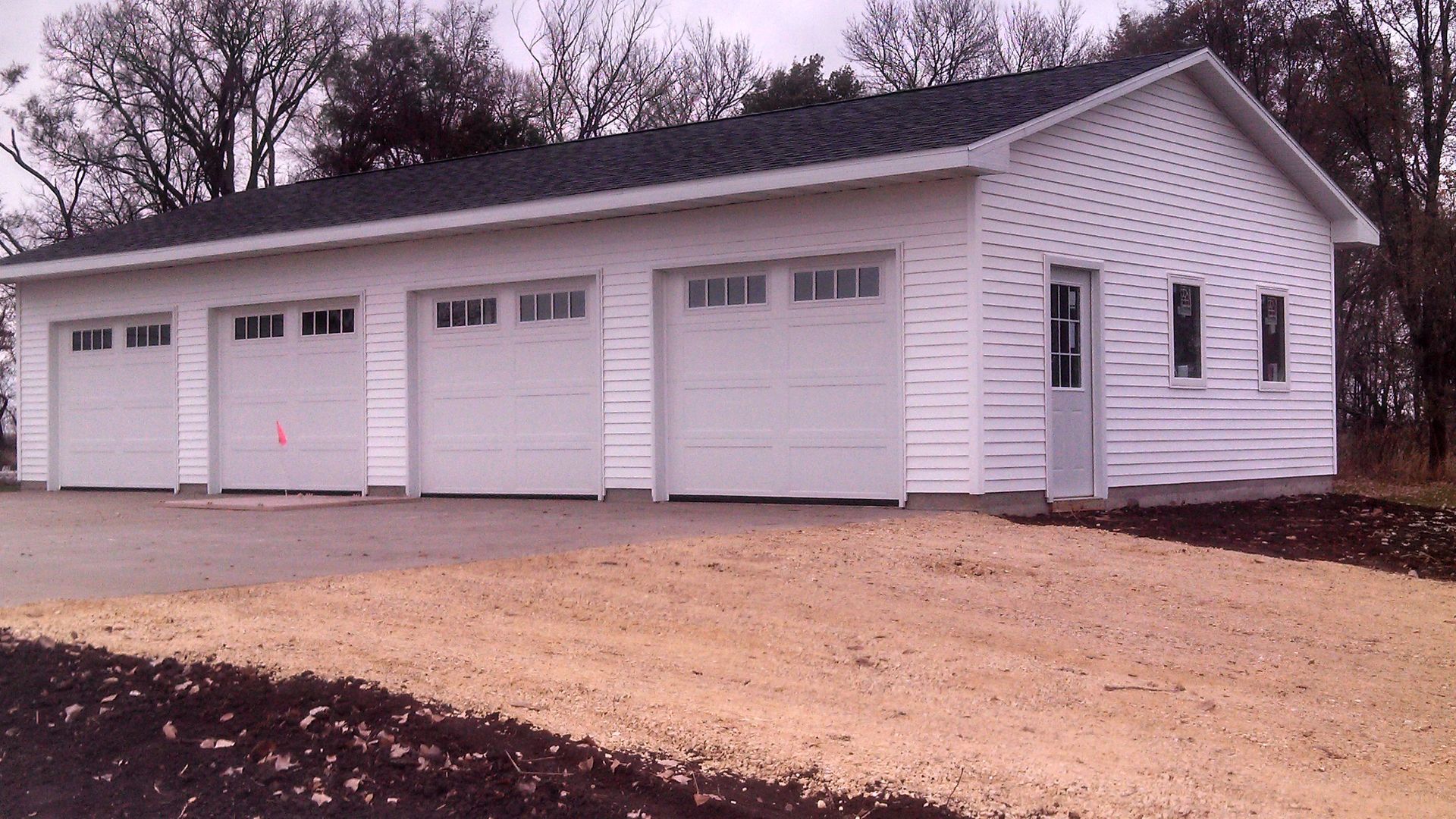White detached four-car garage with gabled roof and paved driveway, set beside trees in a rural setting.