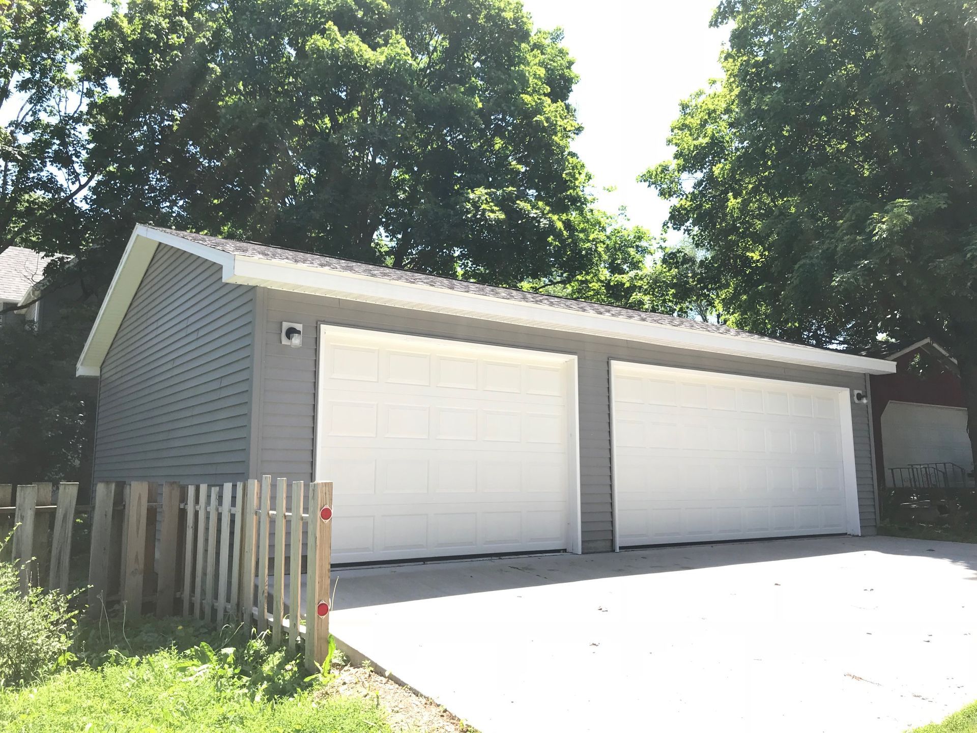 Two-car garage with white doors, gray siding, and a wooden fence under trees