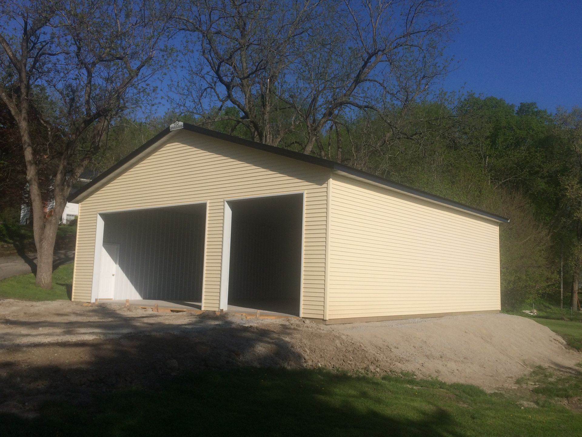 Two-bay beige garage with open doors on a dirt driveway, surrounded by trees and hills.
