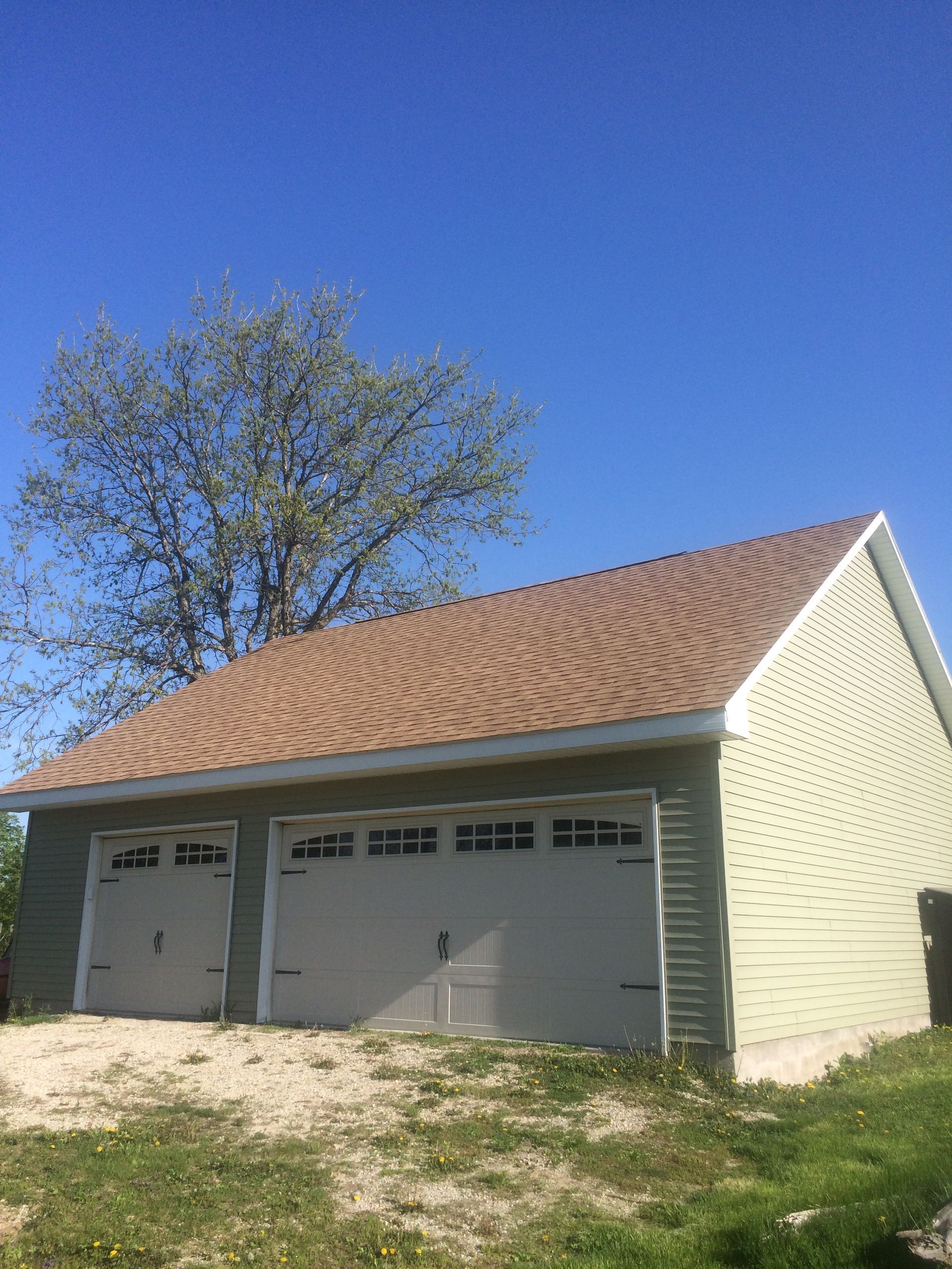 Detached two-car garage with tan siding, brown shingle roof, and a tree beside it under a clear blue sky