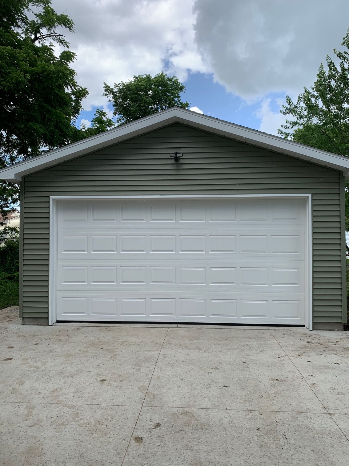 Gray garage with a white door, surrounded by trees and a gravel driveway under a cloudy sky