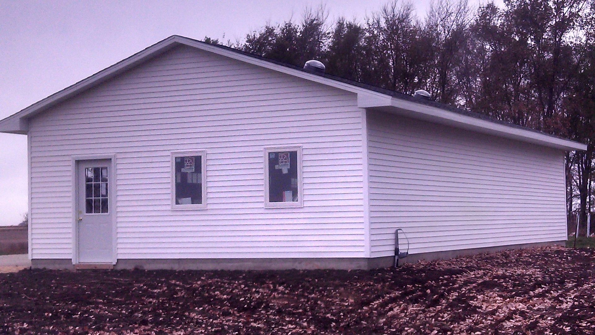 White building with a sloped roof and three windows, standing in a wooded area with leaf-covered ground.