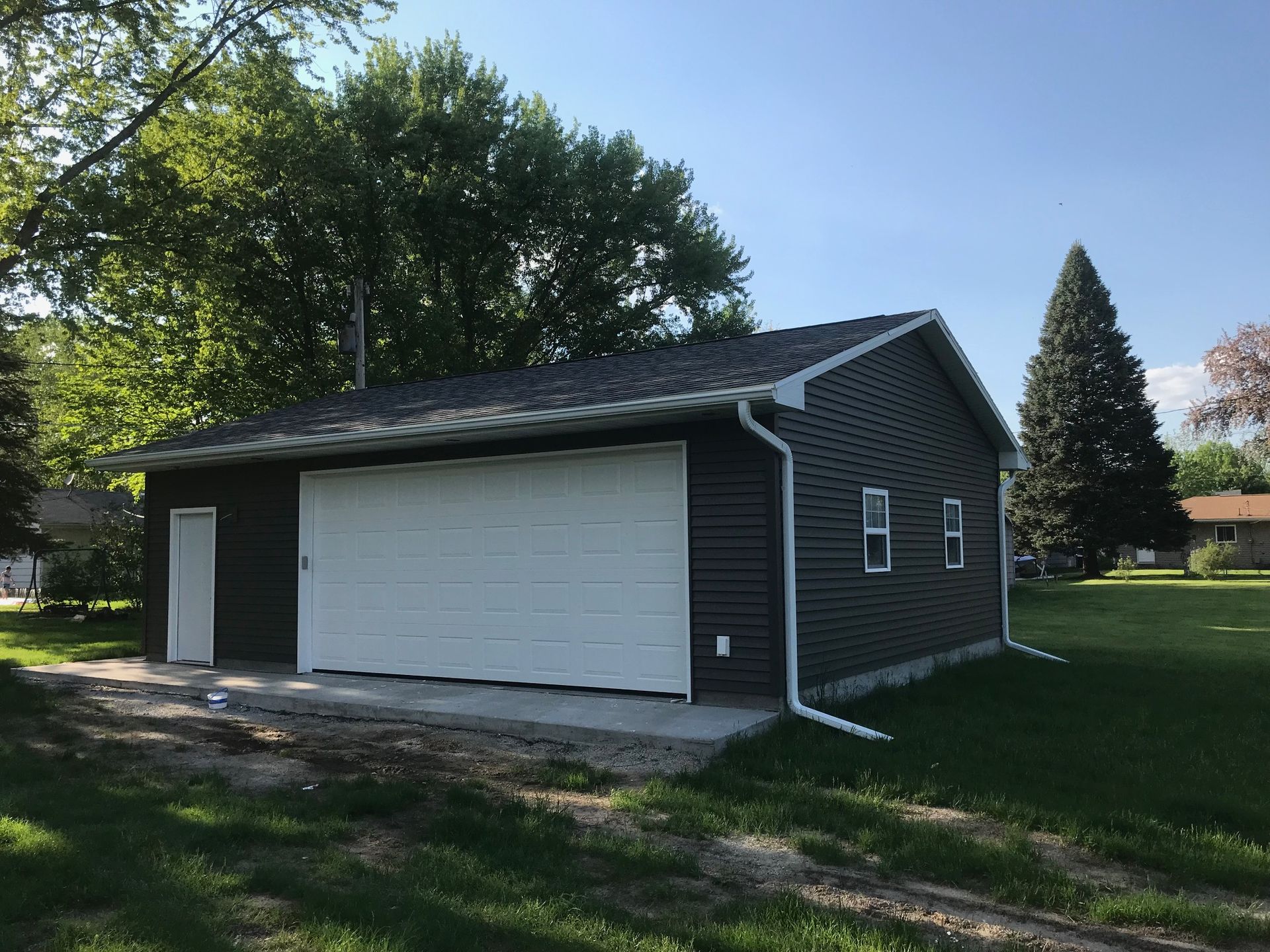 Detached dark gray garage with white doors on a grassy lot, trees and a house in the background