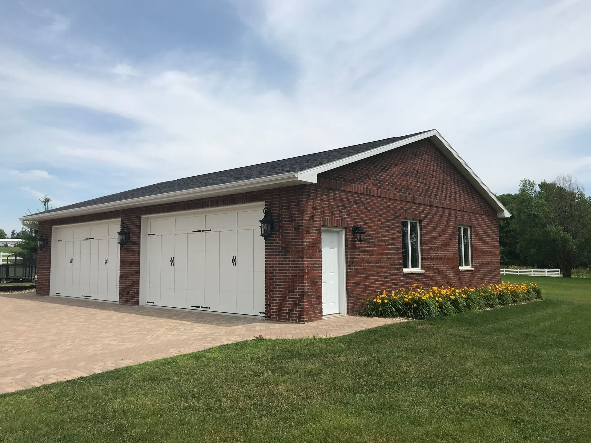Red brick garage with two white doors beside a grassy lawn and flower bed under a cloudy sky