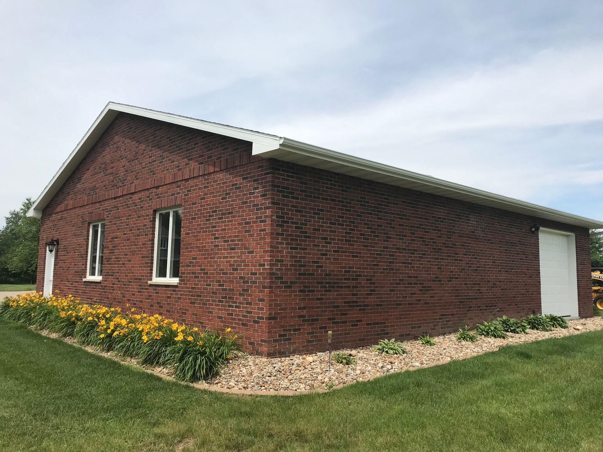 Red brick building with white trim and a metal roof, viewed from the side beside a grassy flower bed.