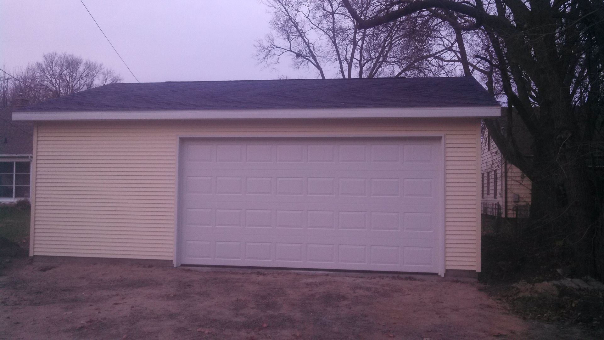 Detached beige garage with a white door at dusk, framed by leafless trees and a bare yard.