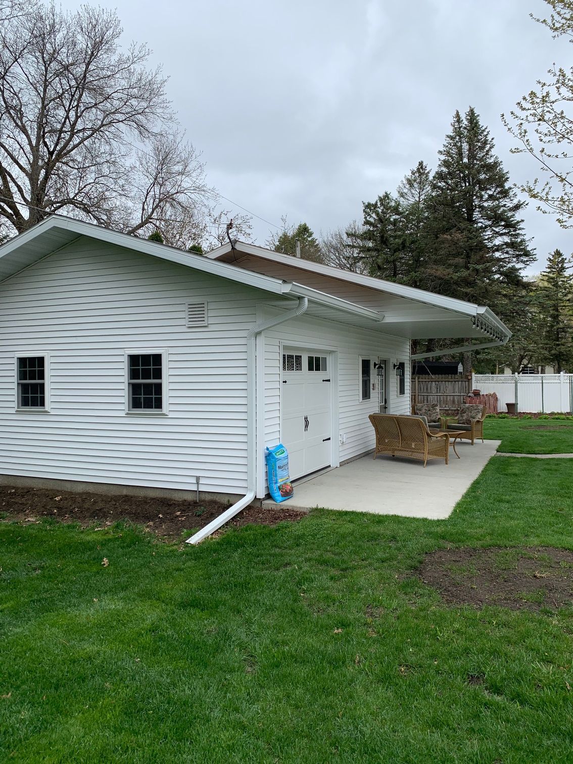 White building with a small porch and stacked firewood, surrounded by green grass and trees