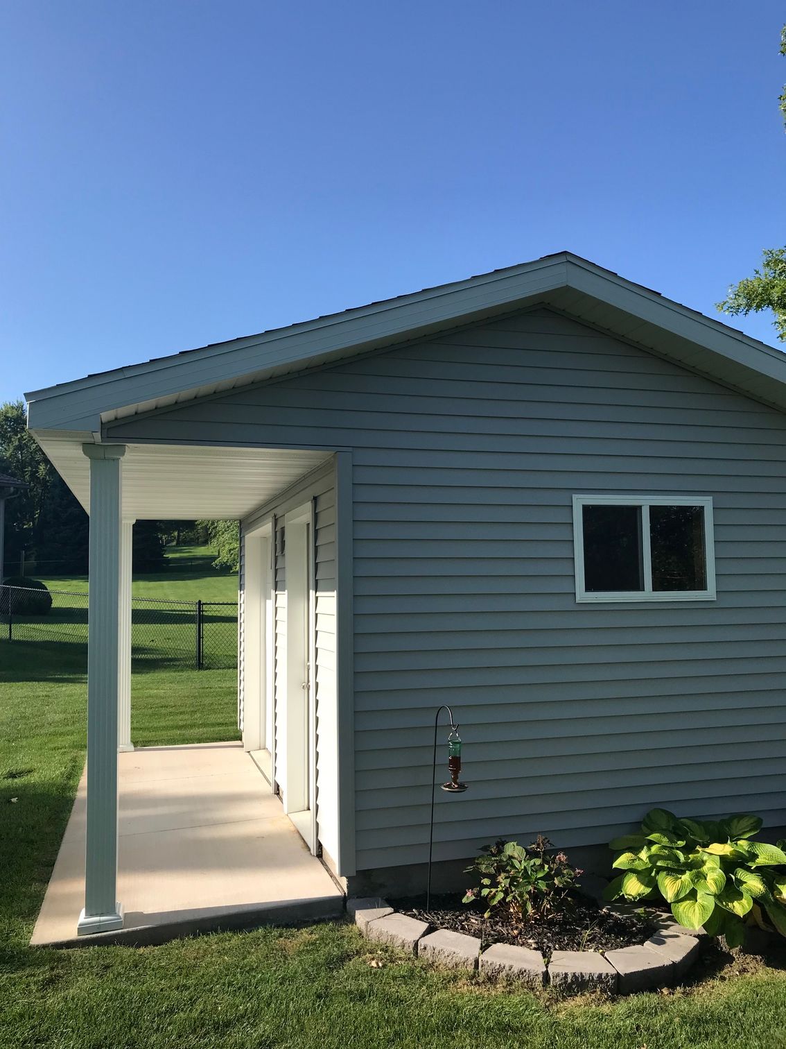 Small gray house side entrance with white porch posts, window, and landscaped bed under a blue sky.