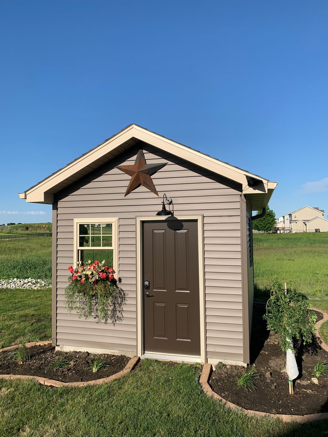 Small tan shed with brown door, window box flowers, and star decor in a grassy field under blue sky