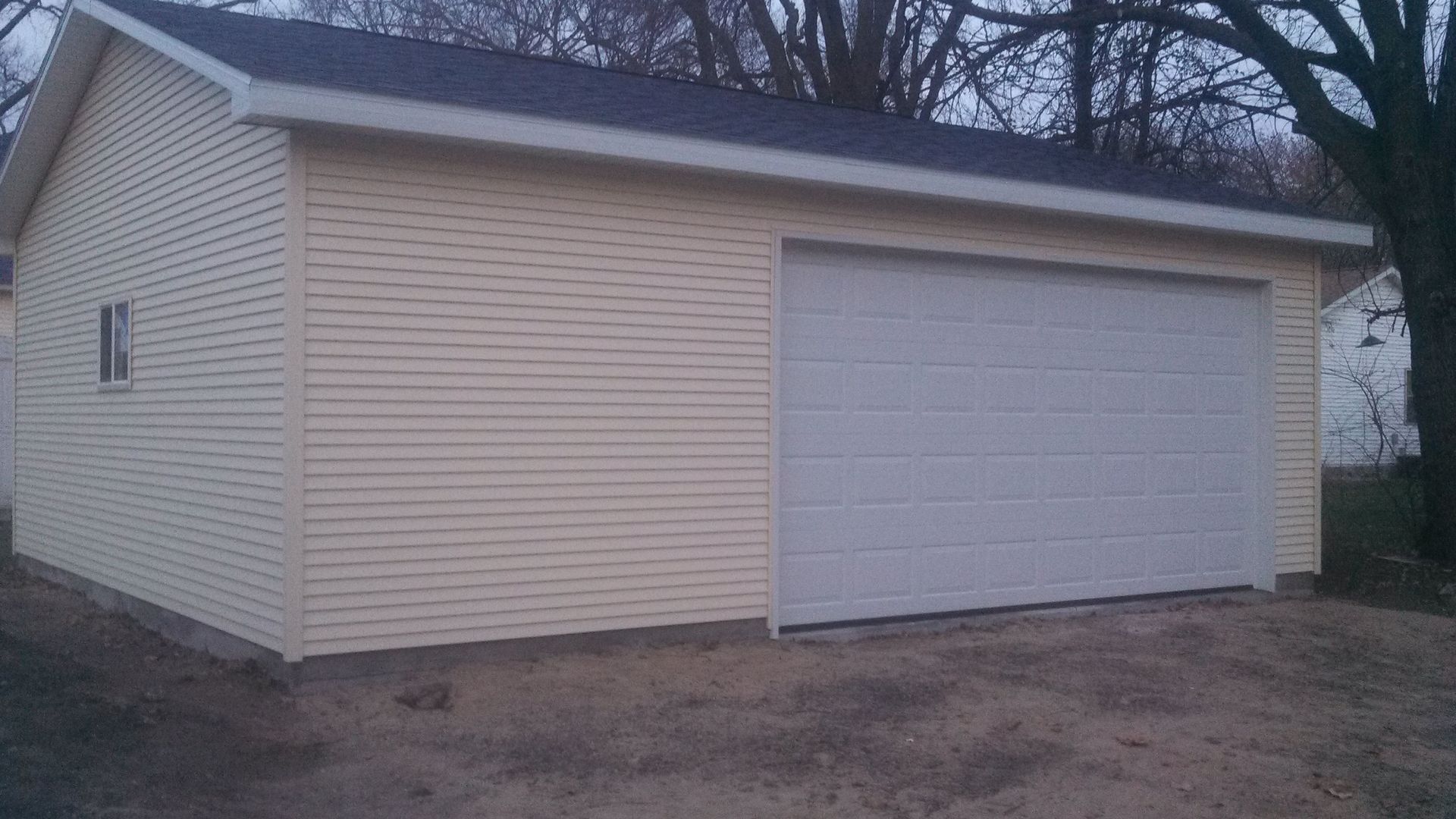 Detached beige garage with a white door and gray shingled roof in a yard.