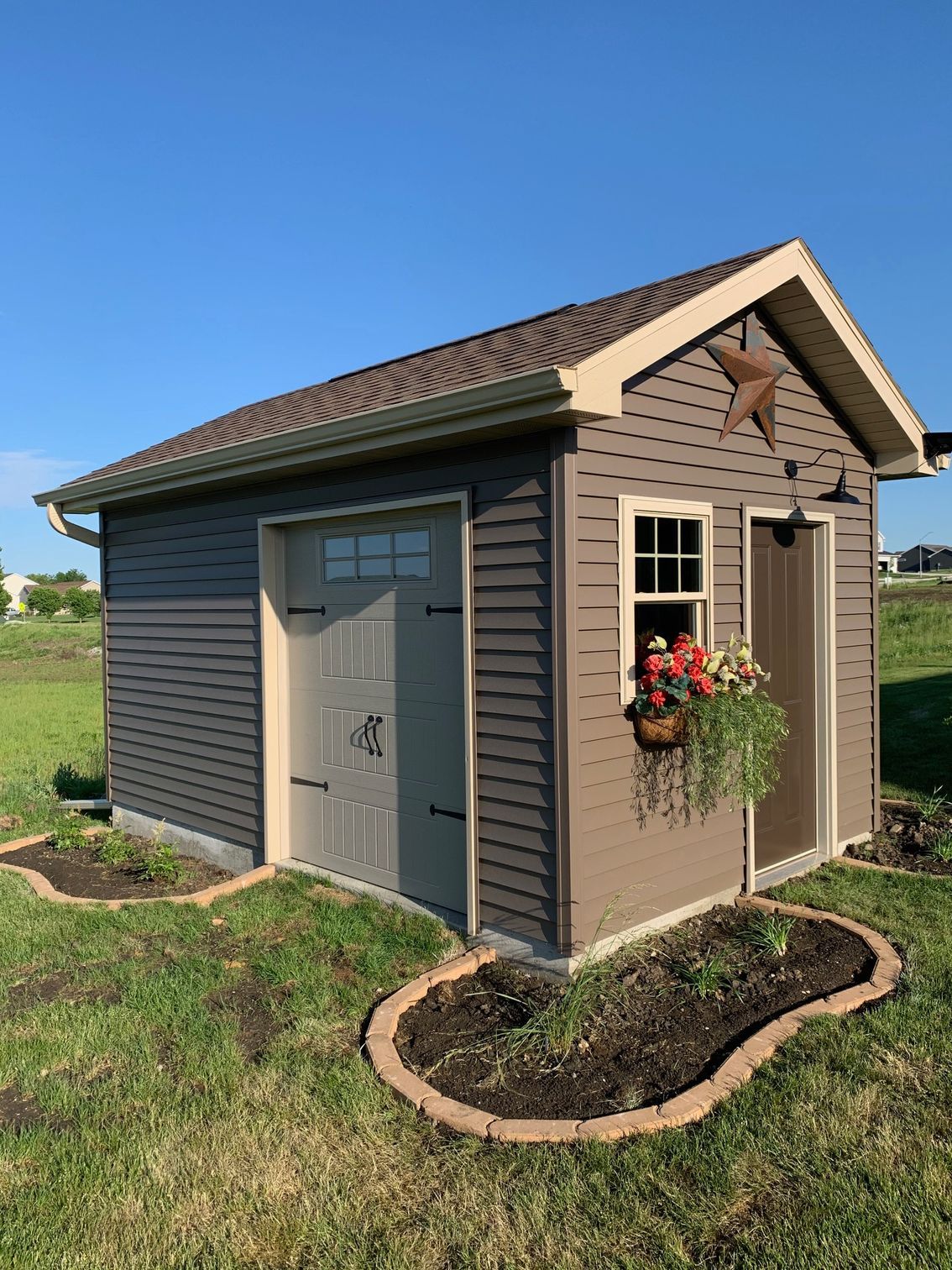 Small brown garden shed with tan trim, white door, window, flower basket, and curved stone border in a grassy yard