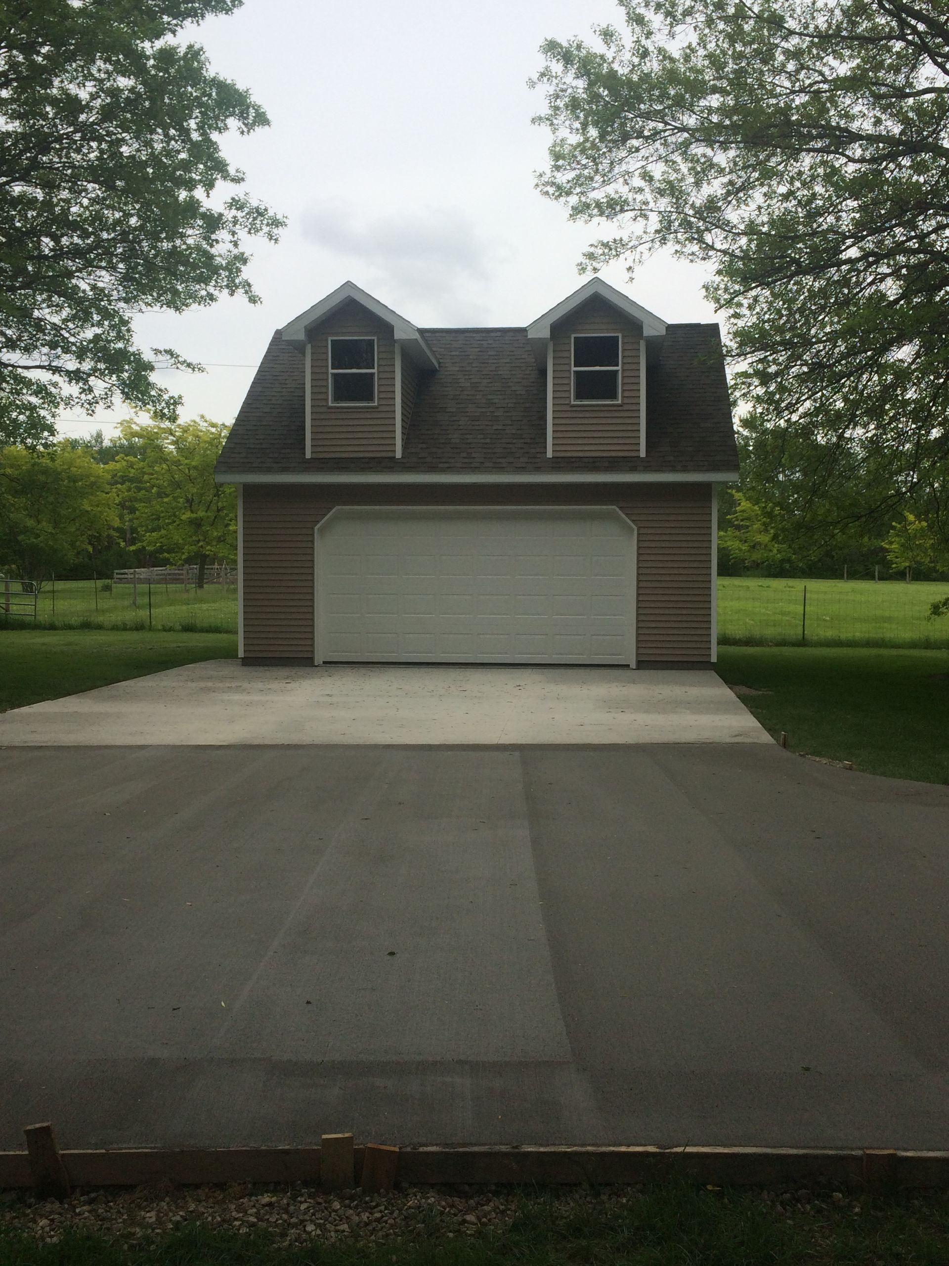 Detached garage with white door and two dormer windows, viewed from a driveway, surrounded by trees and lawn